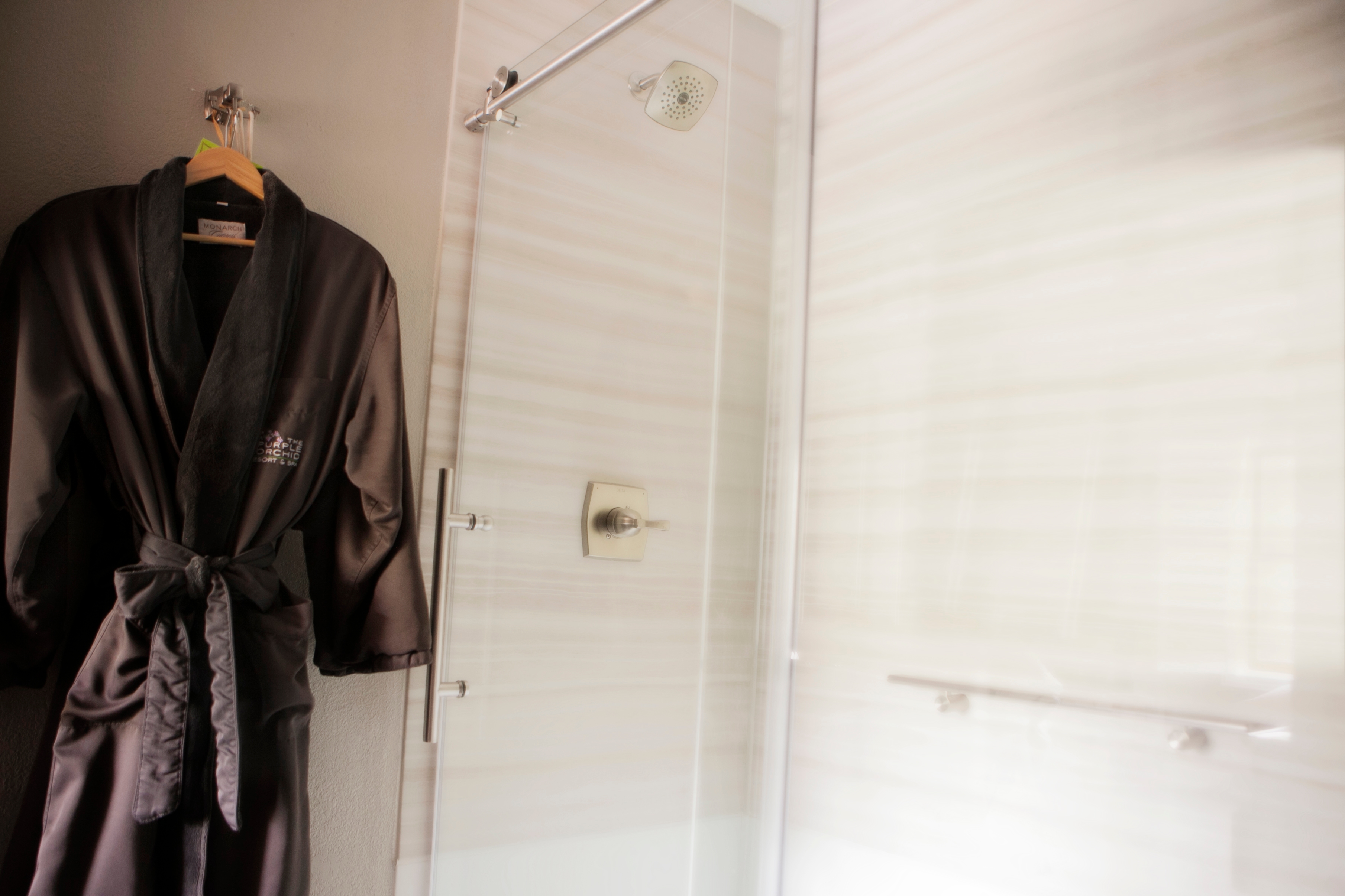 A soft, dark-colored bathrobe hangs on a hook in a modern bathroom. A glass shower door and white tile walls are visible in the background.