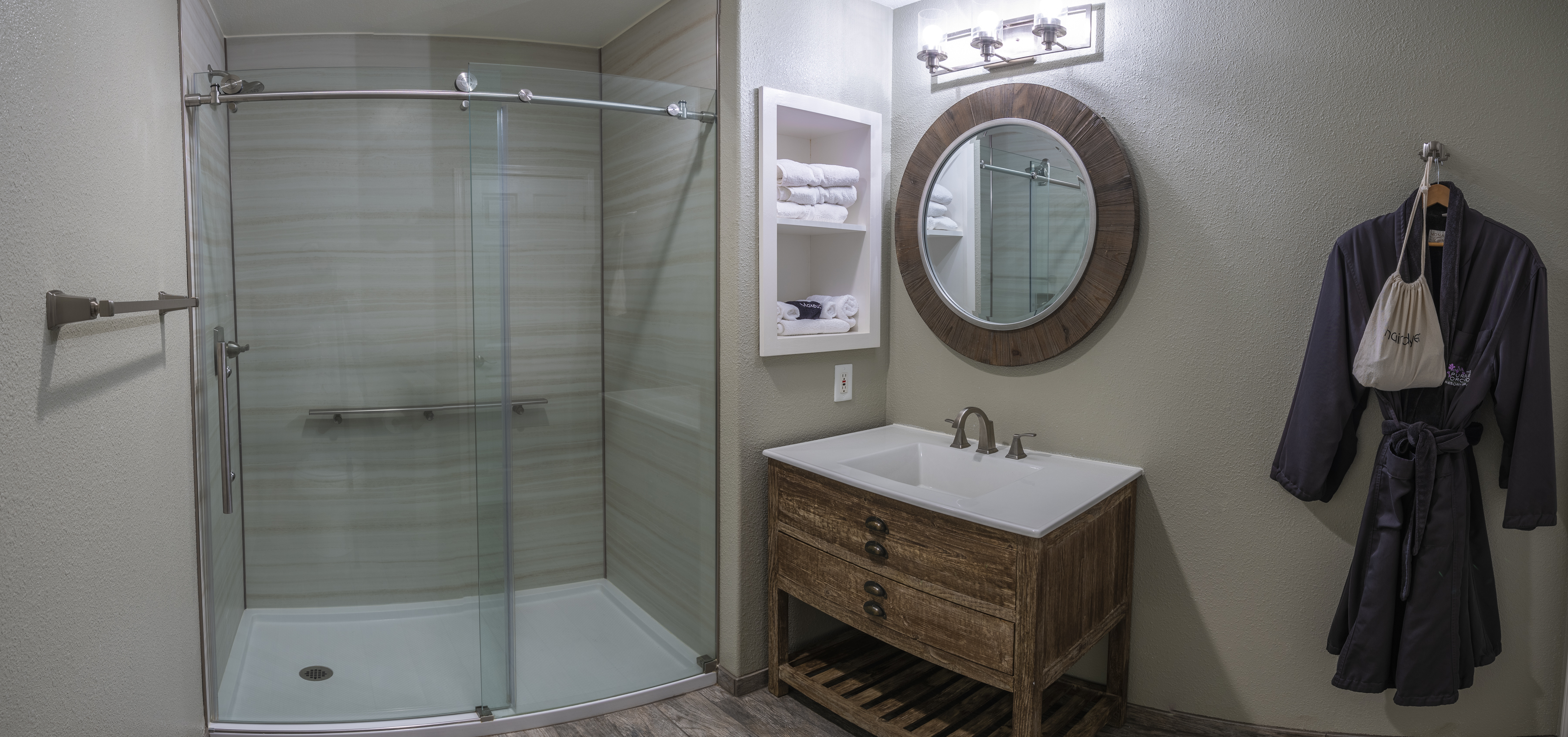 A modern bathroom featuring a walk-in shower with a glass door, a vanity with a sink and a round mirror, and a towel rack. A plush bathrobe hangs on a hook ready for use.