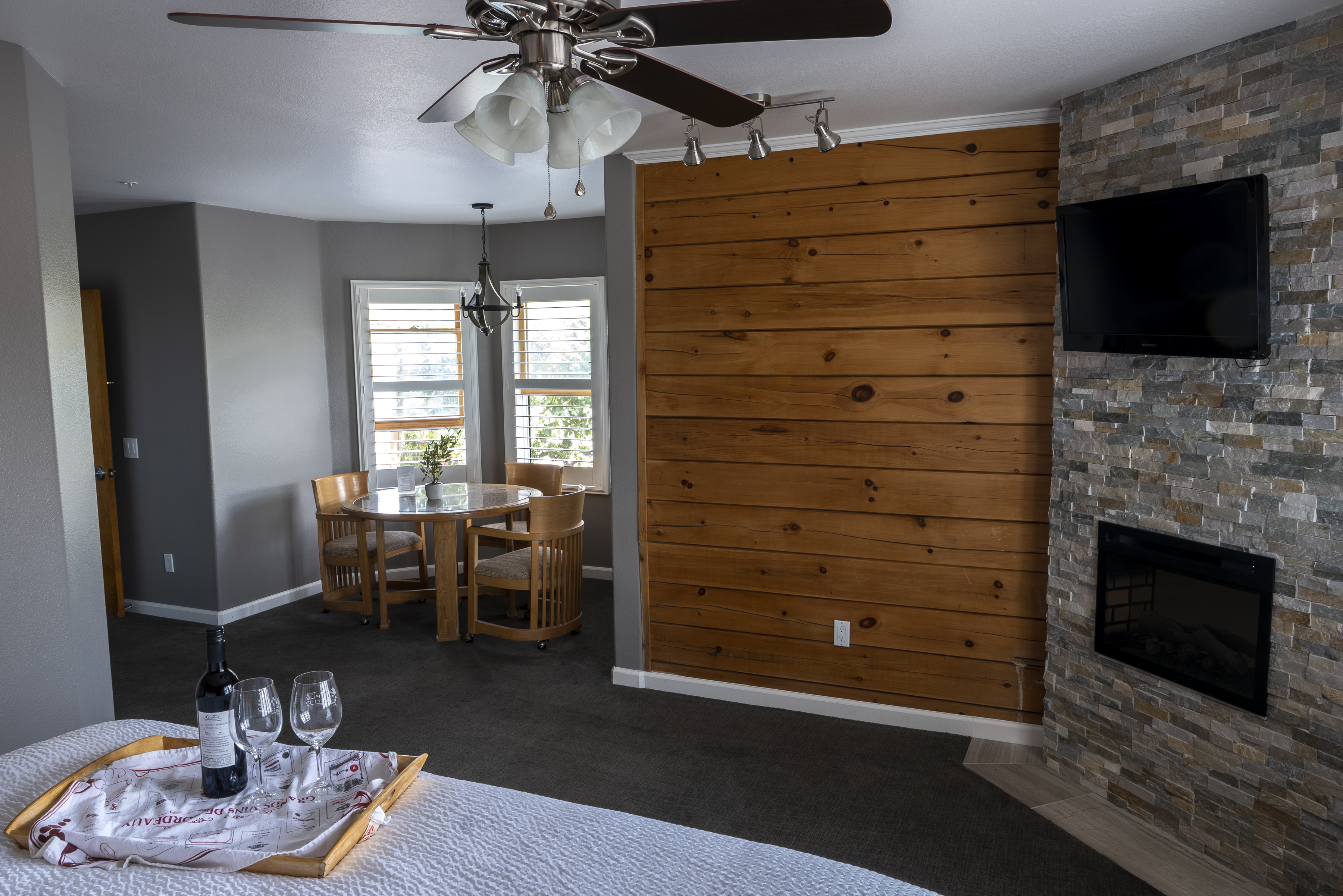 A spacious room with a king-size bed, sitting nook with dining table and chairs, and a fireplace with a stone surround. A wall is adorned with wood paneling, and a ceiling fan hangs overhead.