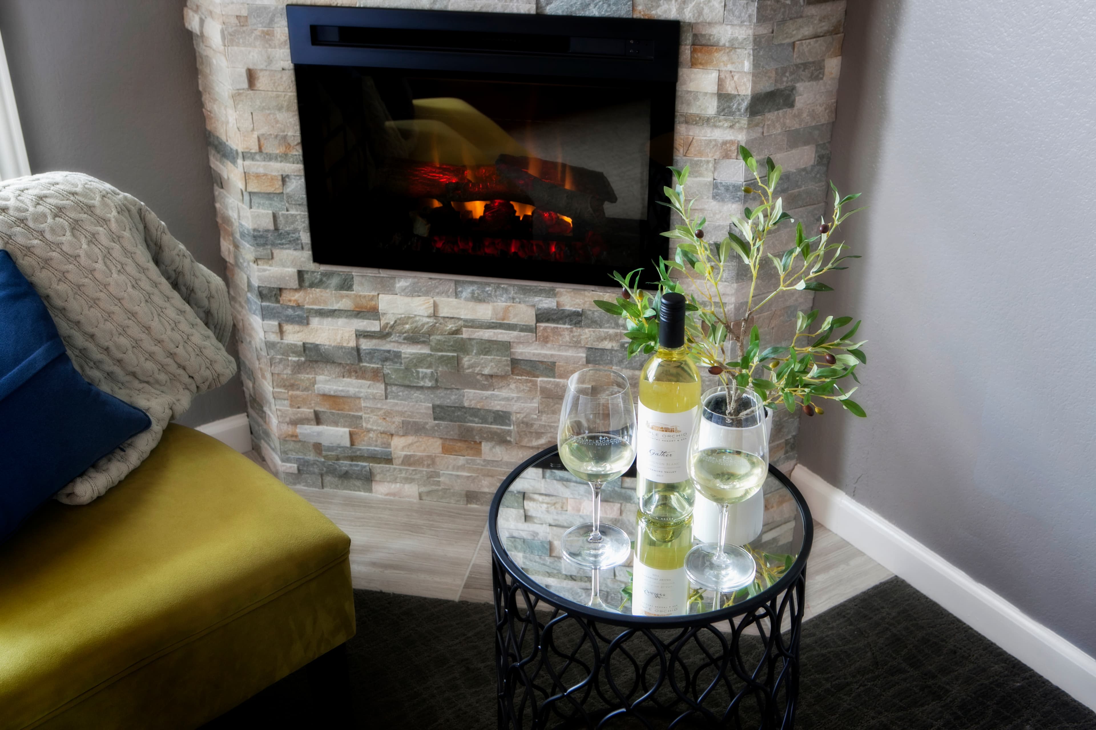 A fireplace with a glowing fire, a small glass-topped table holding a bottle of white wine and two glasses, and a comfortable armchair with a blue throw blanket.