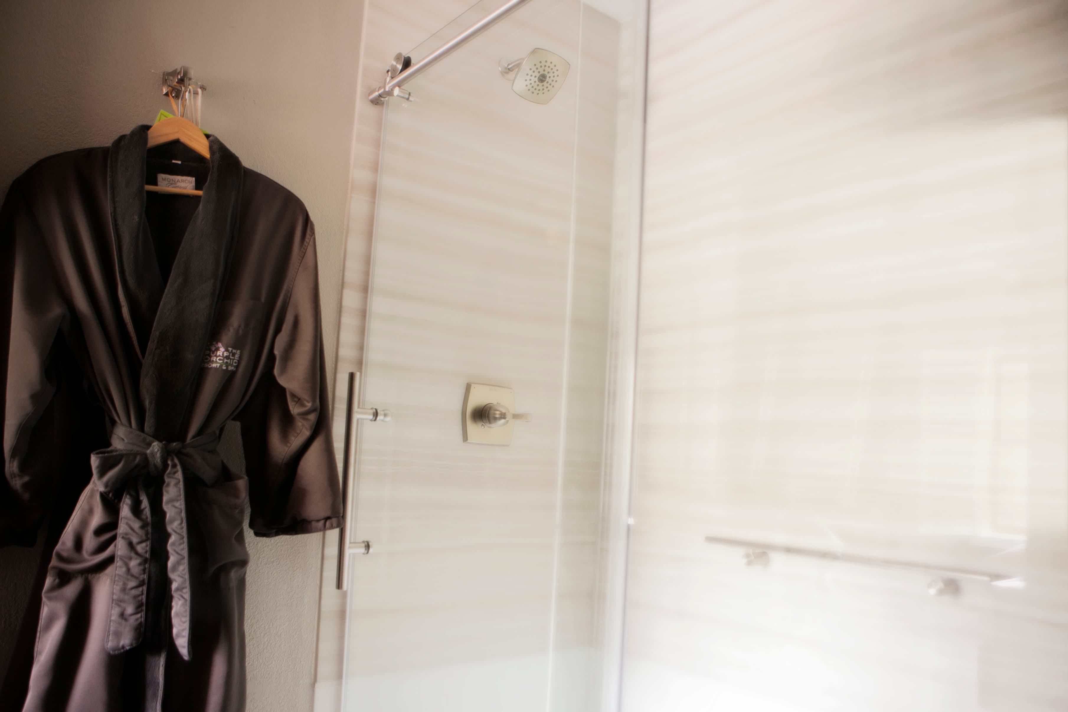 A soft, dark-colored bathrobe hangs on a hook in a modern bathroom. A glass shower door and white tile walls are visible in the background.