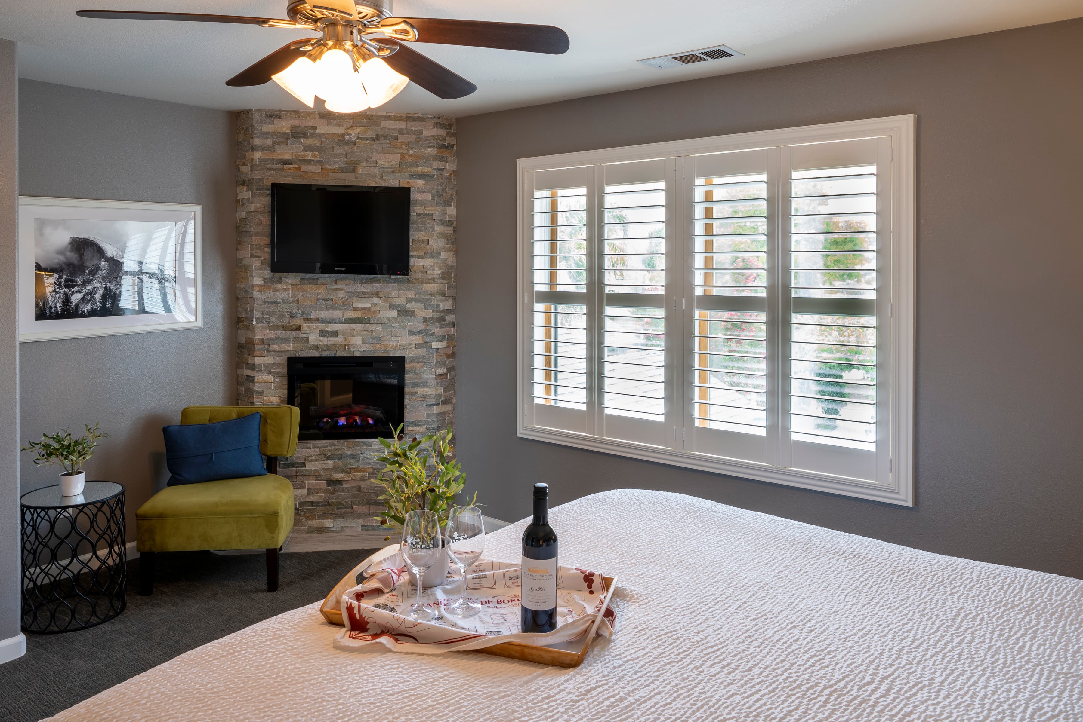 A cozy hotel room with a king-size bed dressed in crisp white linens. A corner fireplace with a stone surround and a seating area with a yellow armchair and a small table. A large, sunny window overlooks a scenic view.