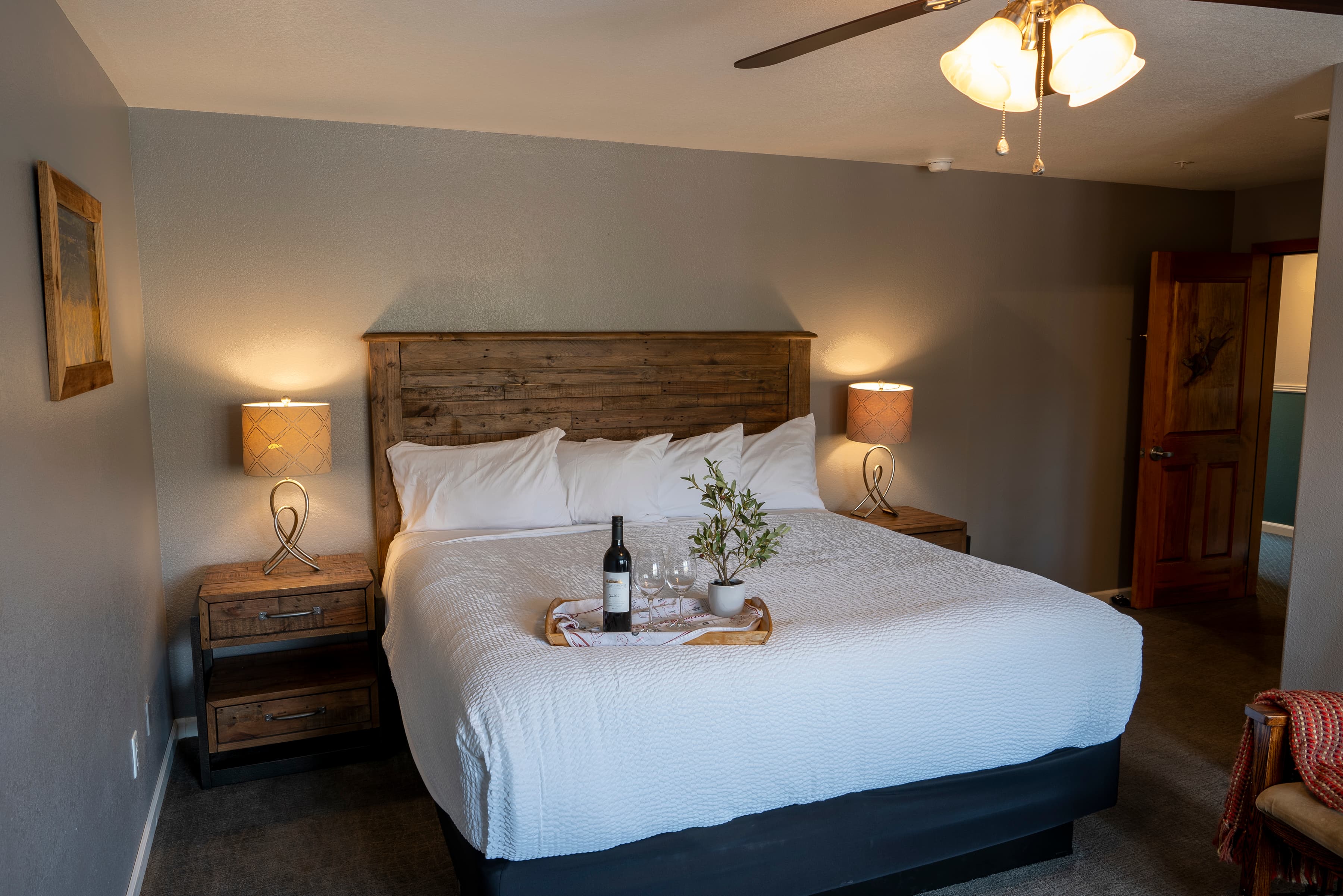 A simply elegant room with a king-size bed dressed in crisp white linens. A wooden headboard and two nightstands with lamps flank the bed. A tray with a bottle of wine and glasses rests on the bed, and a ceiling fan hangs overhead.