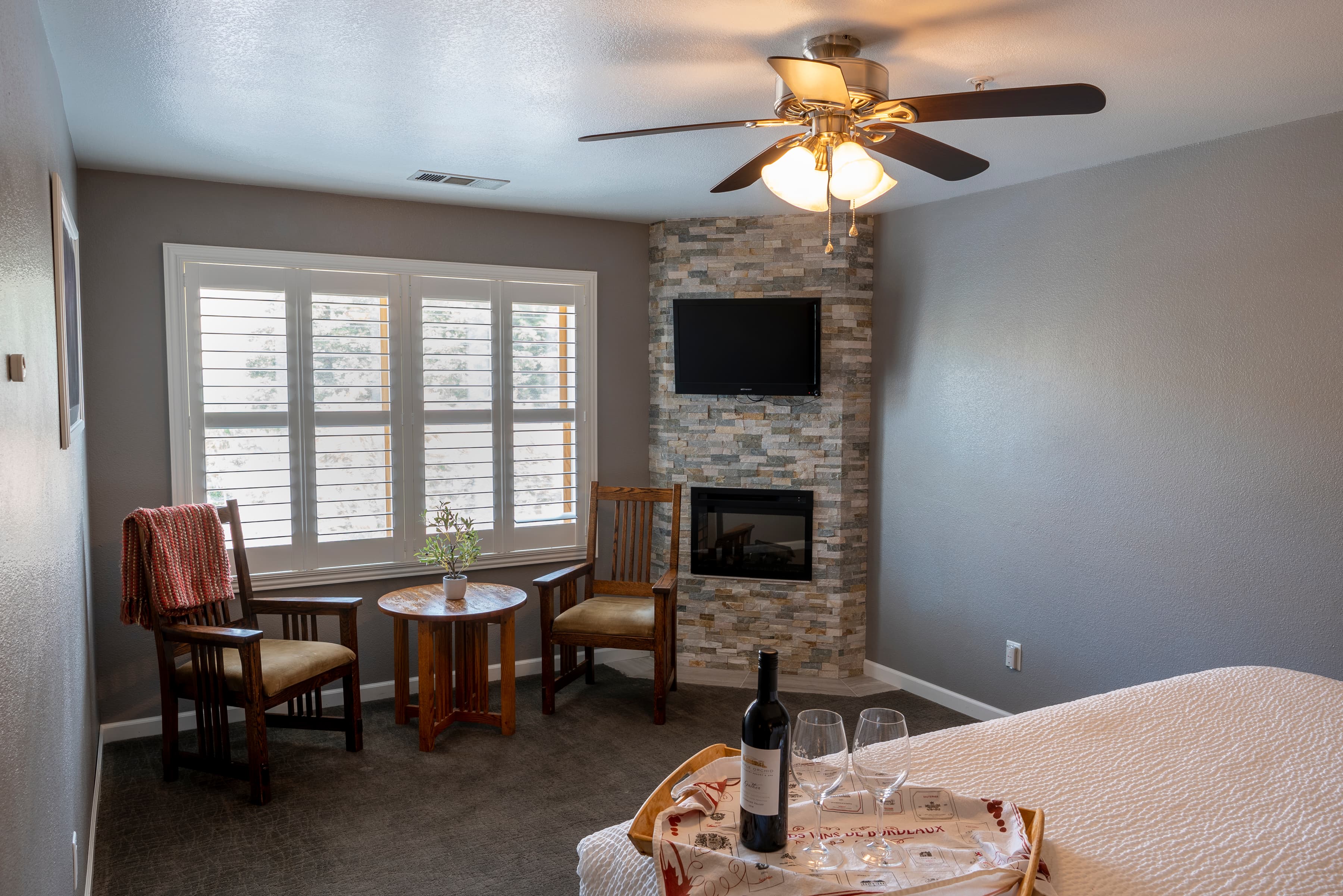An inviting room with a king-size bed, a corner fireplace, two chairs, and a sunny window with white shutters.