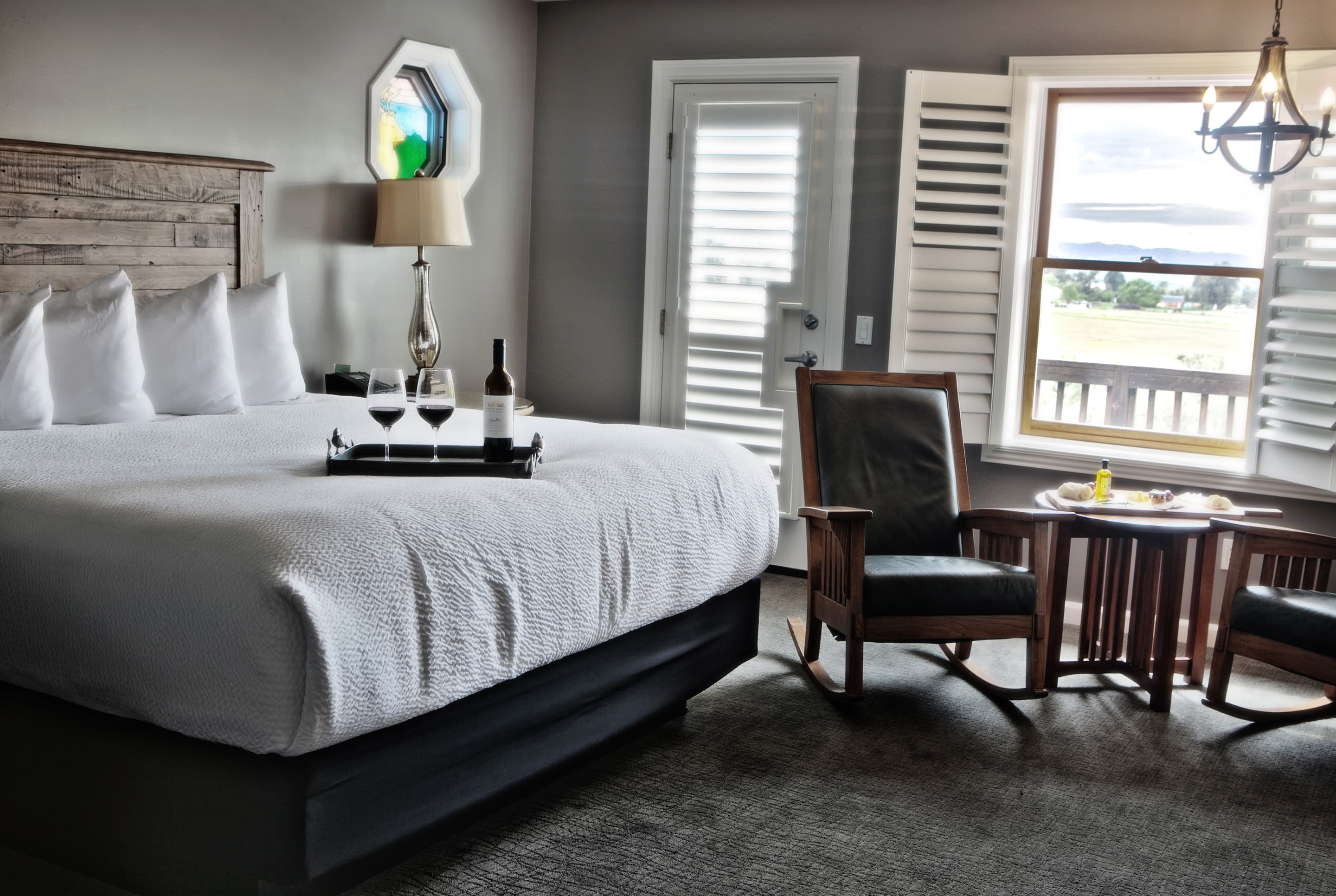 A tranquil room with a king-size bed, two armchairs, a window with shutters, and a tray with a bottle of wine on the bed.