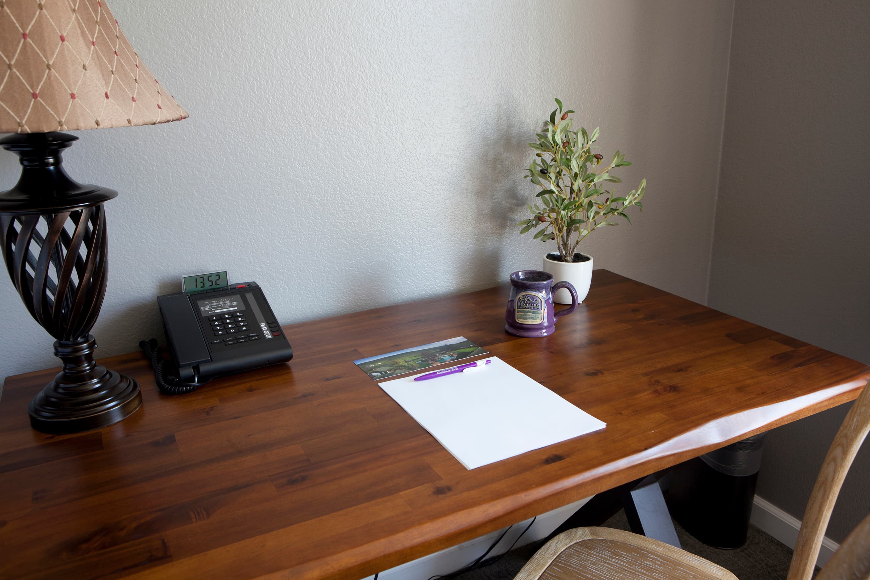 A close-up of a wooden desk with a lamp, a telephone, a small potted plant, and a mug. A sheet of paper and a pen lie on the desk.
