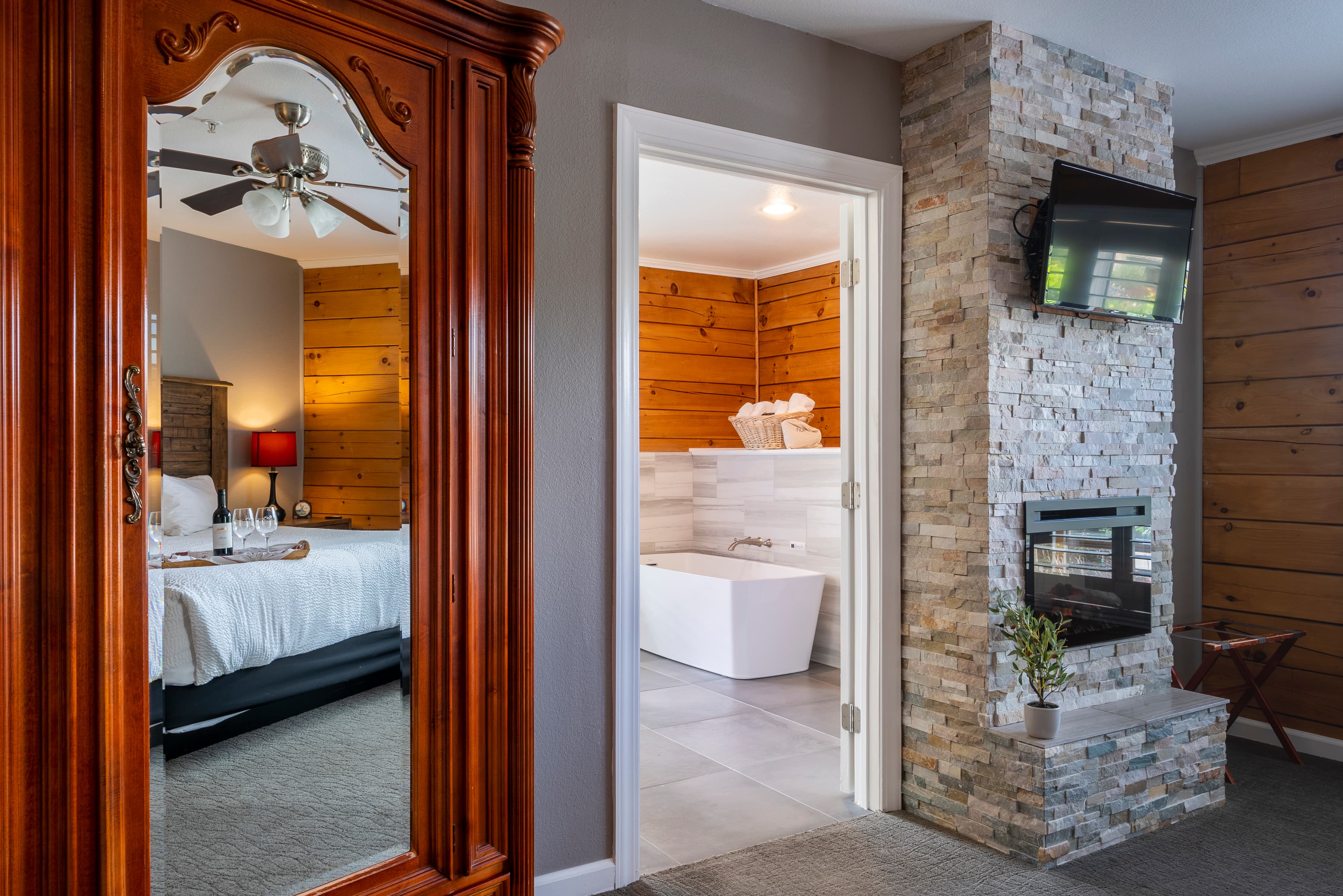 A cozy room with a king-size bed dressed in crisp white linens. A fireplace with a stone surround, and a wooden armoire reflects the bed in its mirror. A doorway leads to a spacious bathroom with a freestanding bathtub.