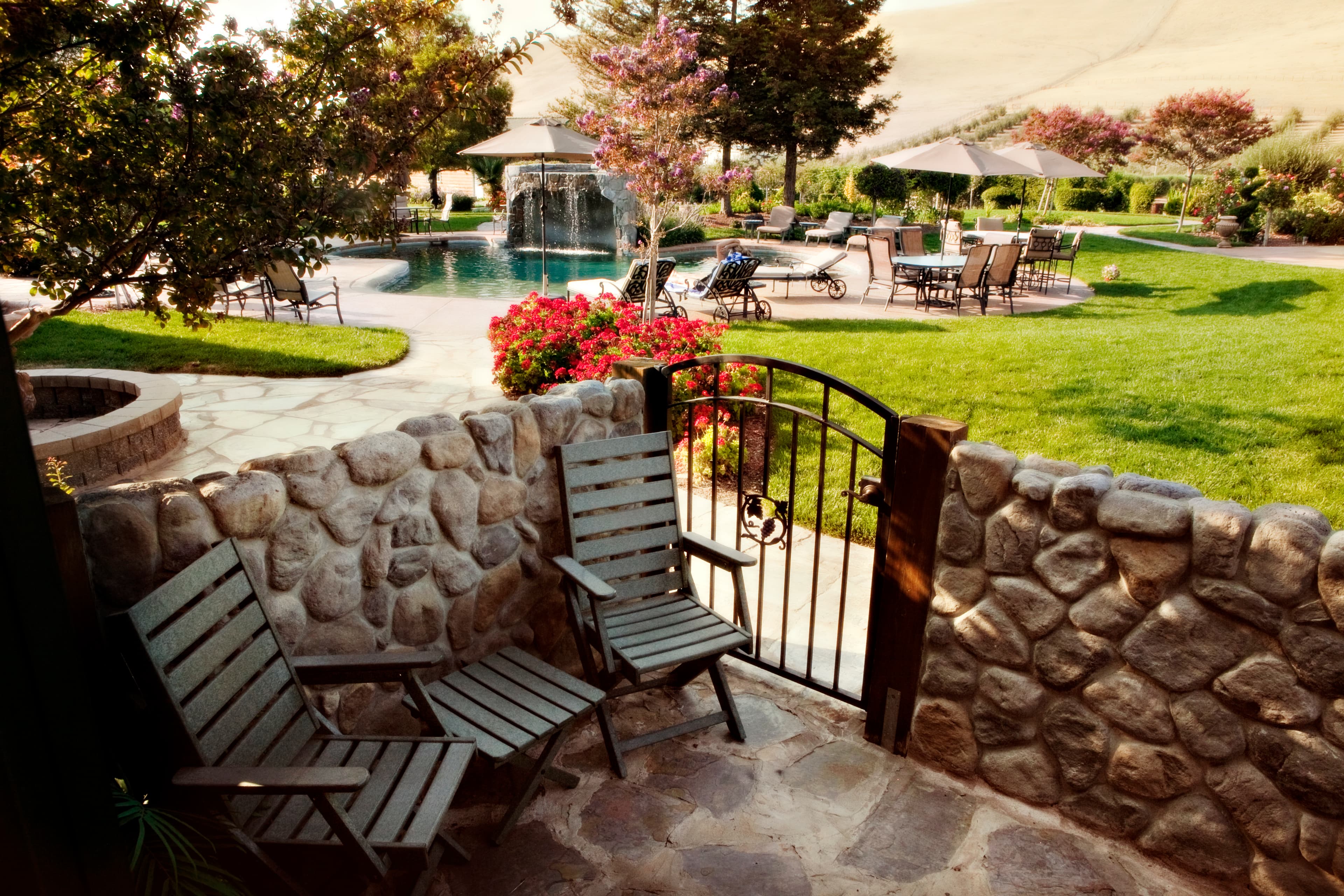 A view from a patio with two wooden chairs. The patio is surrounded by a stone wall and overlooks a beautiful outdoor space with a swimming pool, lounge chairs, and lush green lawns.