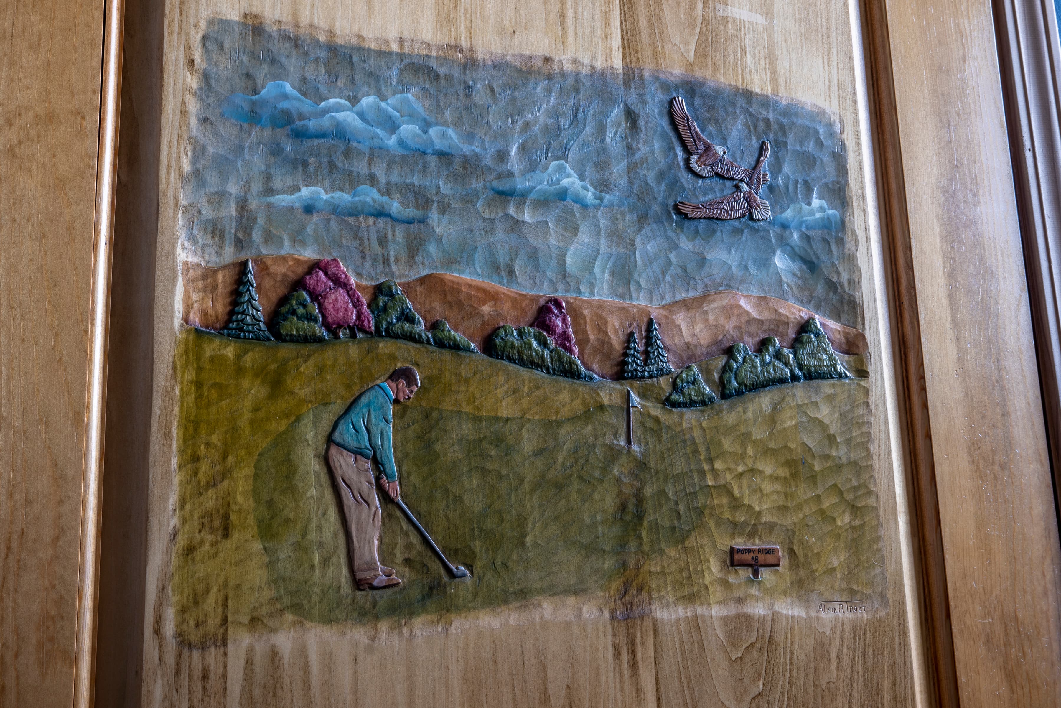 A detailed wood carving depicting a golfer in mid-swing on a golf course. The carving includes a scenic background with hills, trees, and a flying bird.