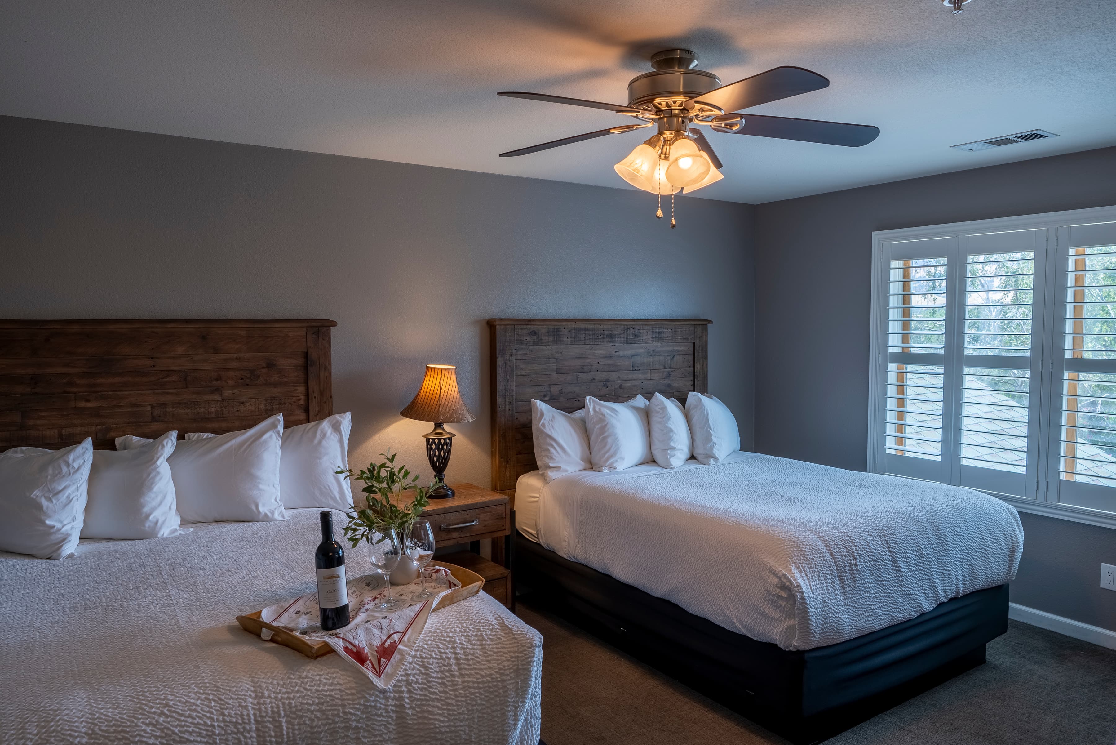 A spacious room with two queen-size beds dressed in crisp white linens. A ceiling fan hangs overhead, and a large window overlooks a scenic orchard view.