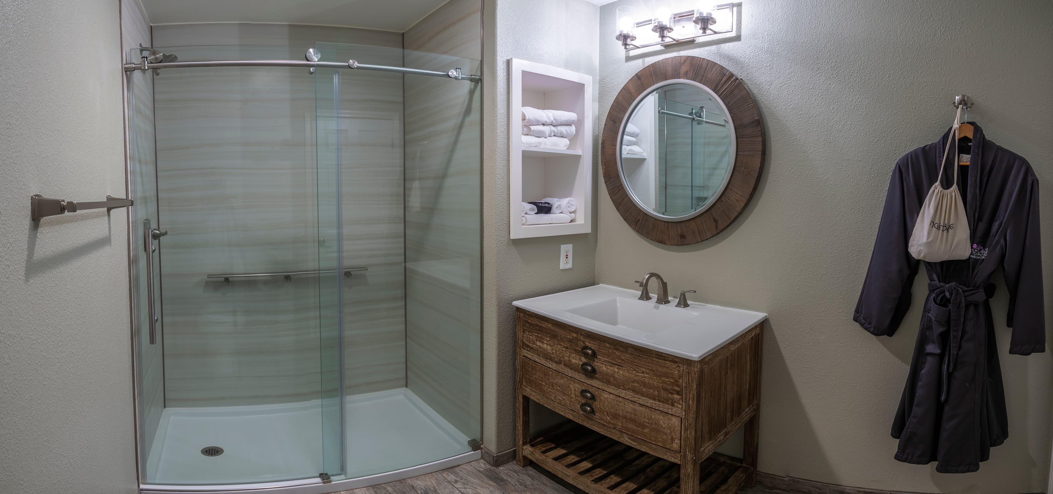 A modern hotel bathroom featuring a walk-in shower with a glass door, a vanity with a sink and a round mirror, and a towel rack. A plush bathrobe hangs on a hook ready for use.