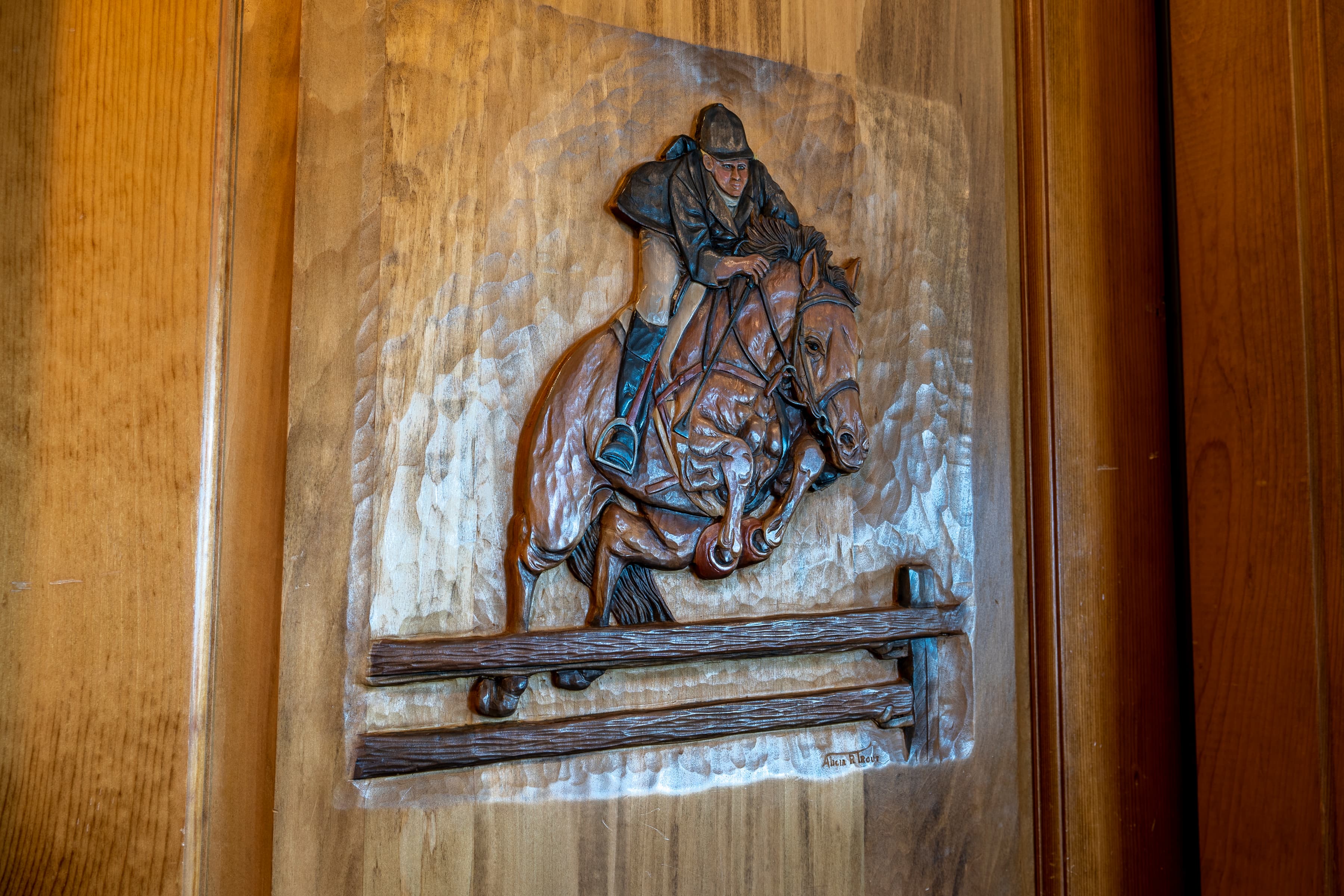 A detailed wood carving depicting a horse and rider in mid-jump over a fence. The carving is done in relief on a wooden surface.