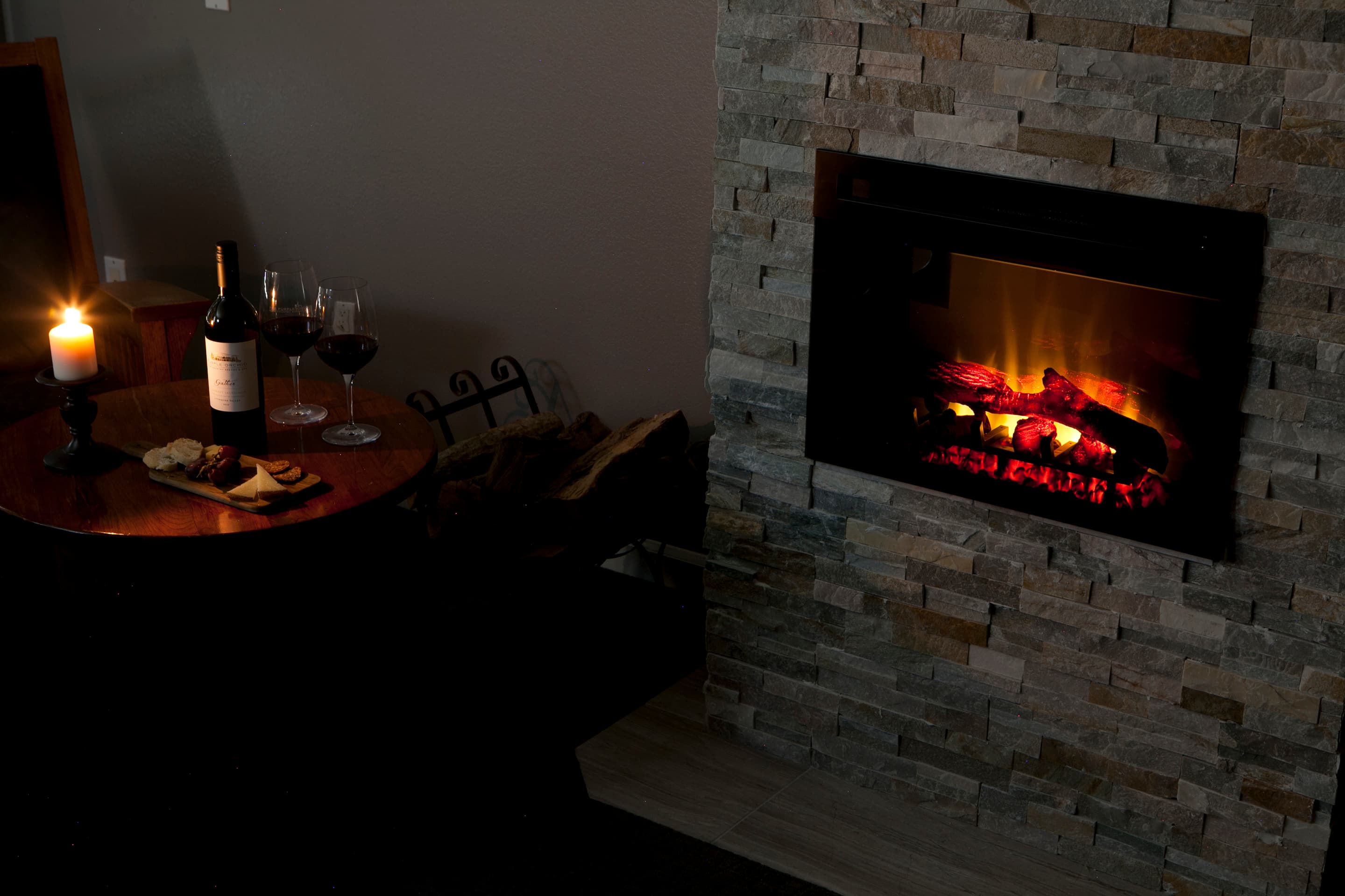 A cozy corner with a fireplace glowing softly. A small table is set with a bottle of wine, glasses, and a candle creating a romantic ambiance.