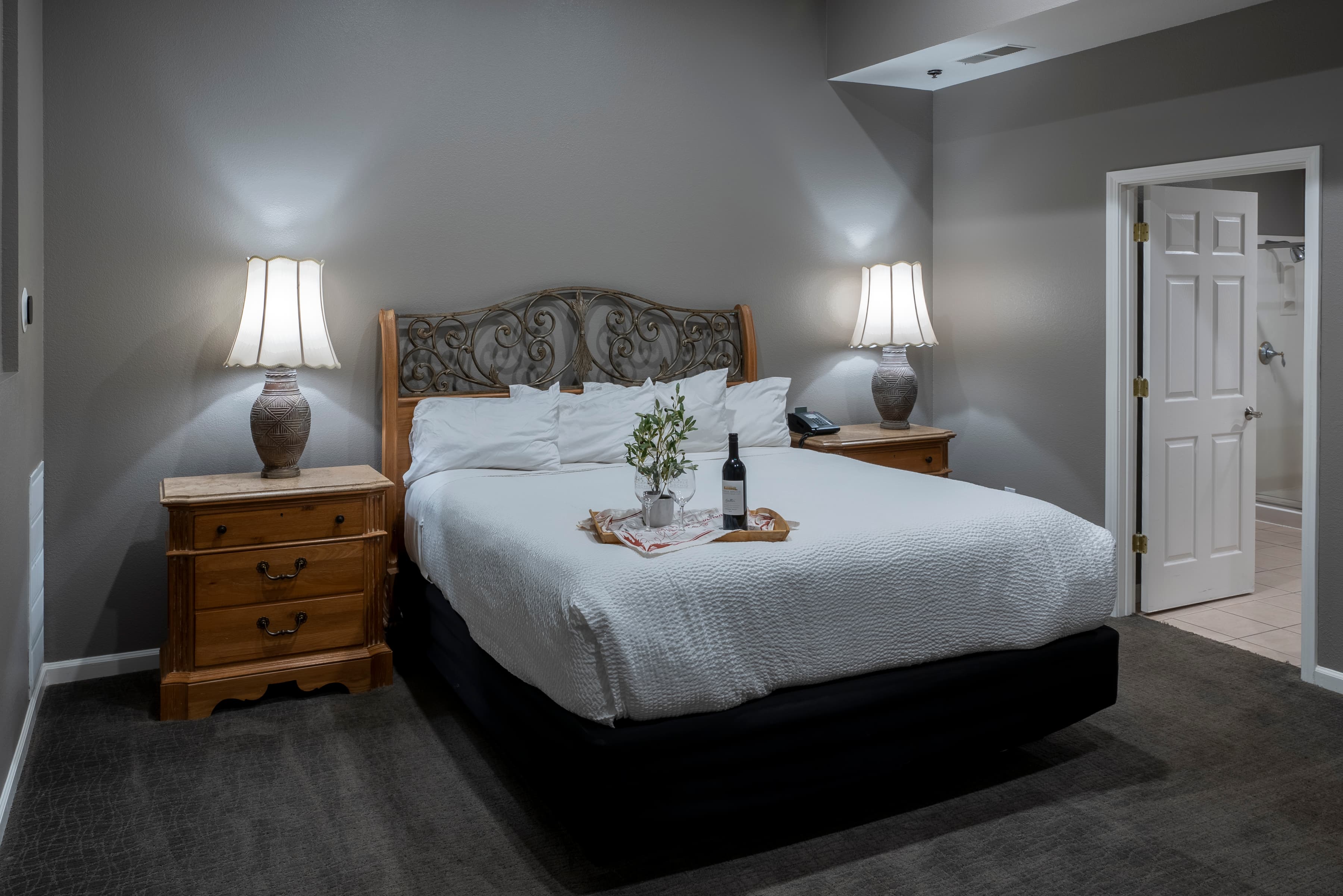 A cozy room with a king-size bed dressed in crisp white linens. Two nightstands with lamps flank the bed, and a tray with a bottle of wine and glasses rests on the bed.