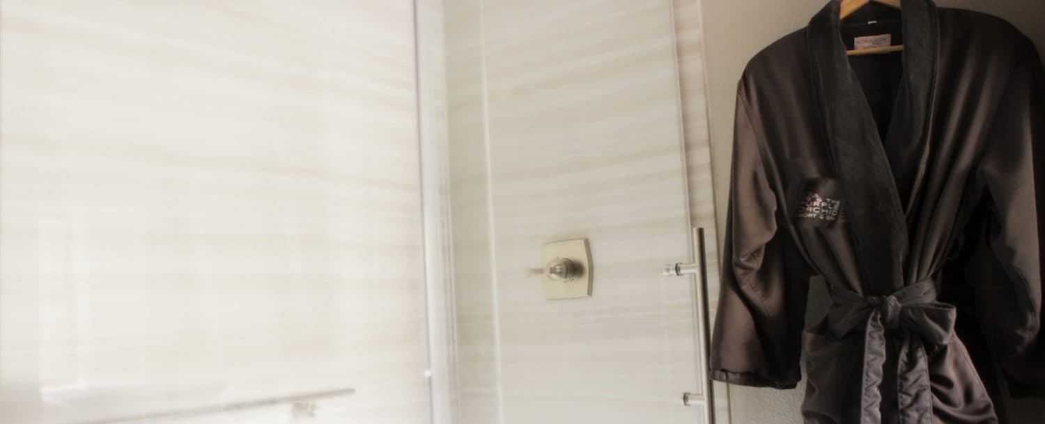 A soft, dark-colored bathrobe hangs on a hook in a modern bathroom. A glass shower door and white tile walls are visible in the background.