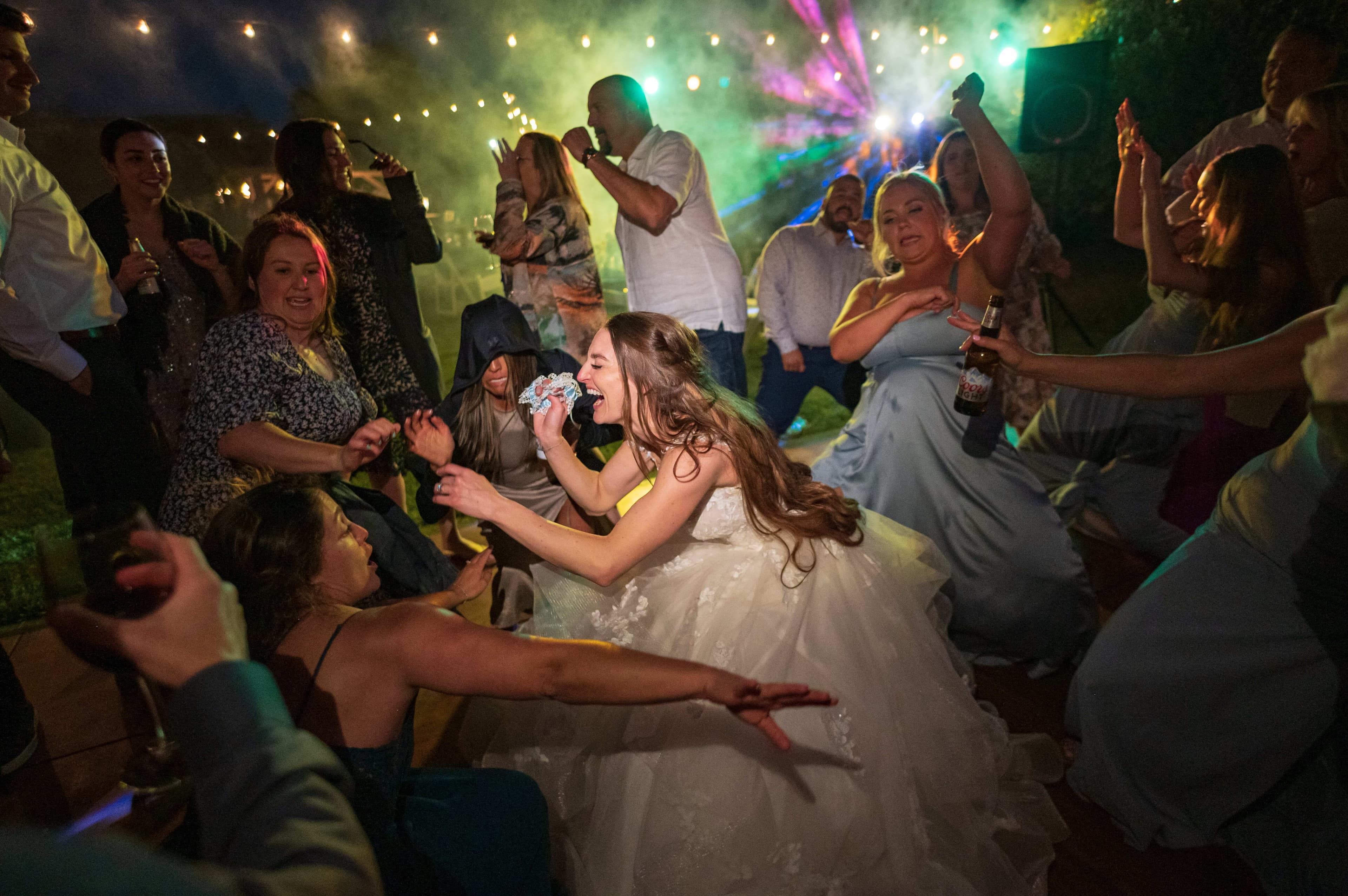 A bride joyfully dances among a lively crowd during a wedding celebration, surrounded by colorful lights and festive energy.