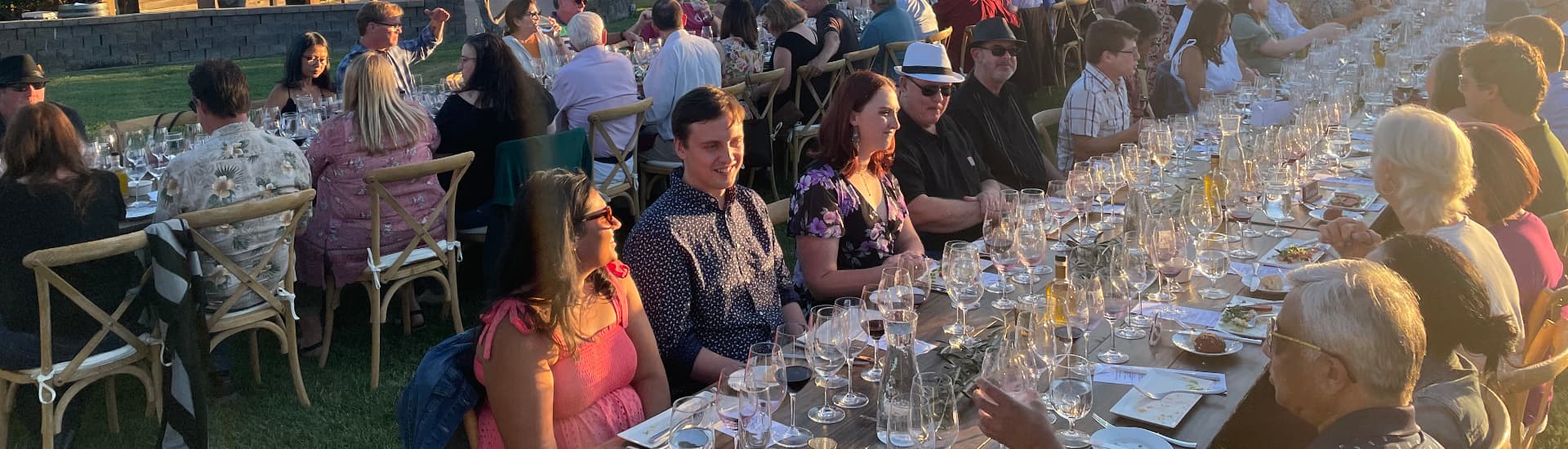 A large group of people enjoys a sunset dinner at a long table set with glasses and plates.