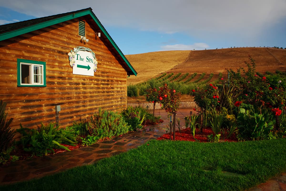 A wooden spa building with a sign pointing left, surrounded by lush gardens and hills in the background. A wooden spa building with a sign pointing left, surrounded by lush gardens and hills in the background.