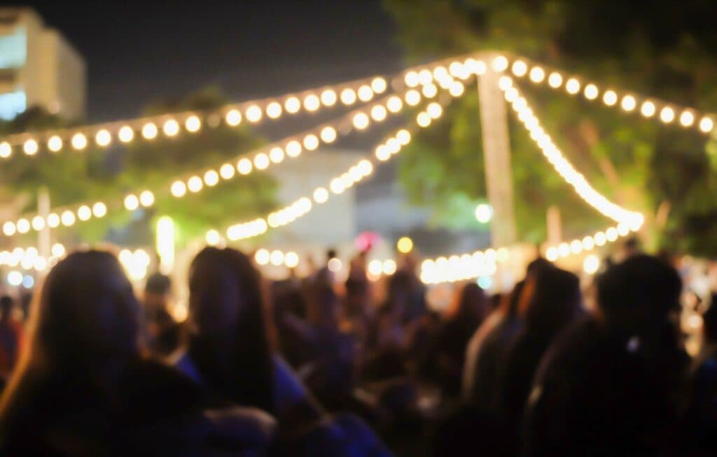 A blurred nighttime scene featuring string lights and people gathered at an outdoor event. A blurred nighttime scene featuring string lights and people gathered at an outdoor event.