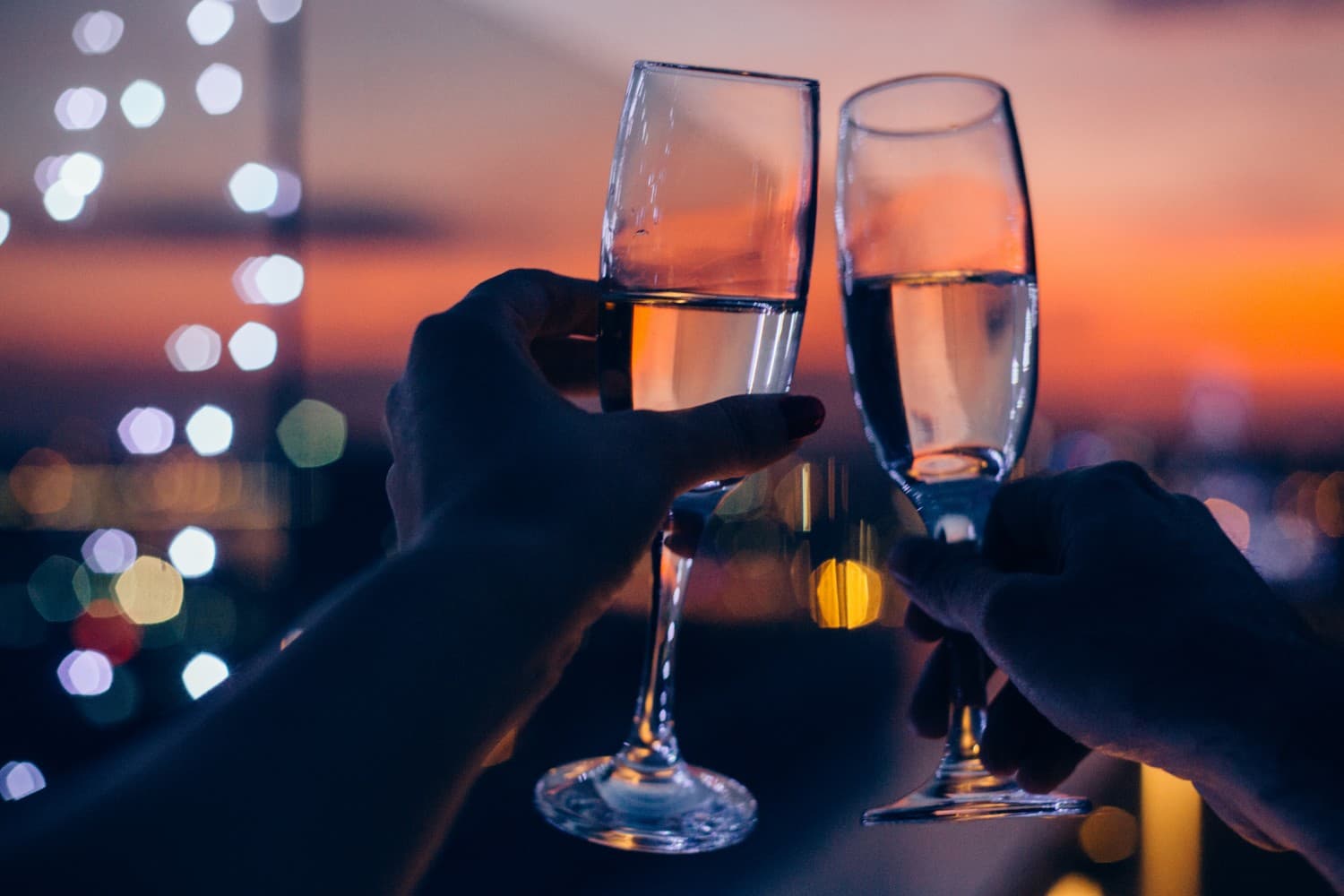 Two hands clinking champagne glasses against a colorful sunset backdrop. Two hands clinking champagne glasses against a colorful sunset backdrop.