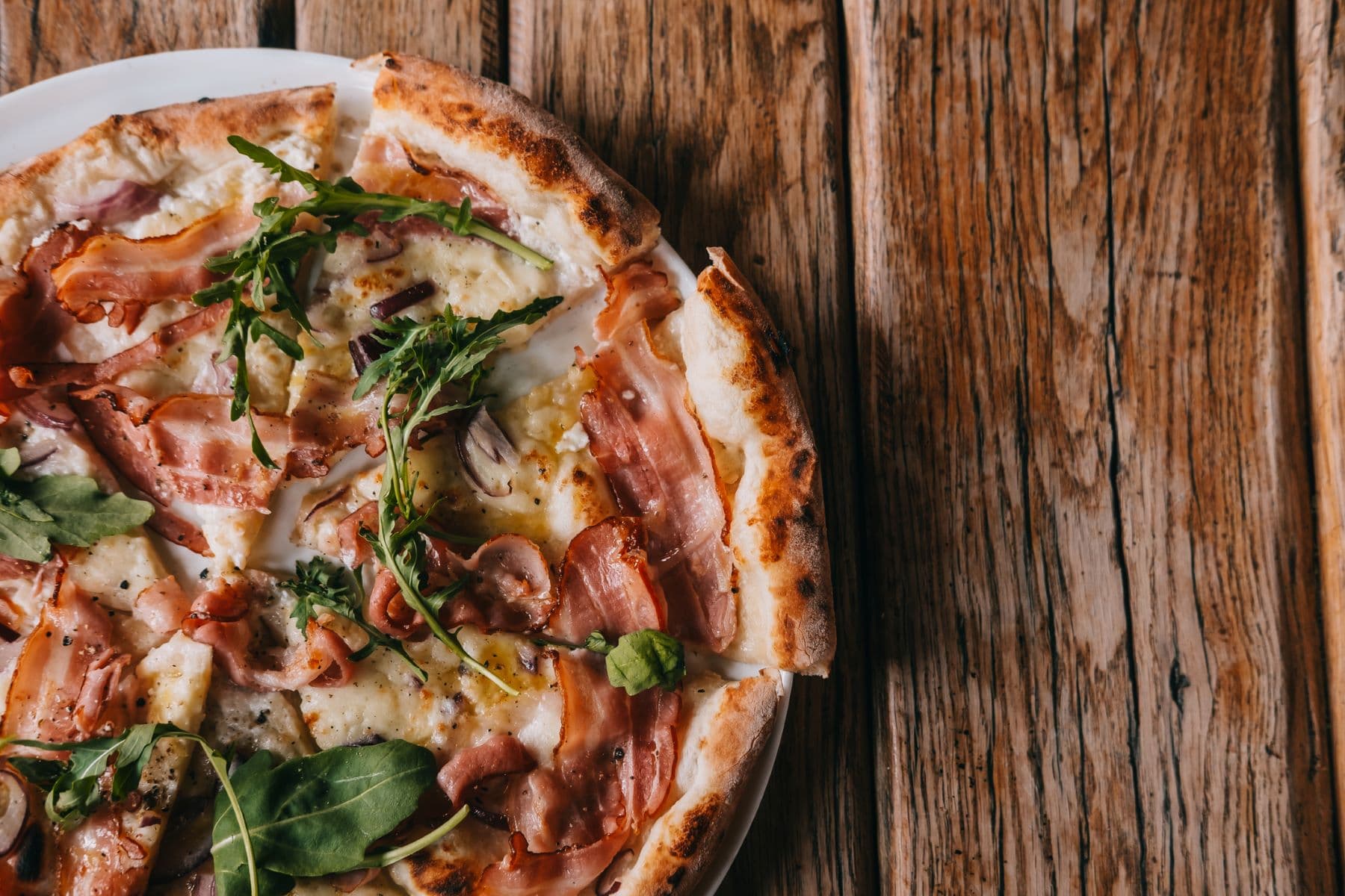 A close-up of a pizza topped with ham, arugula, and cheese on a wooden table. A close-up of a pizza topped with ham, arugula, and cheese on a wooden table.