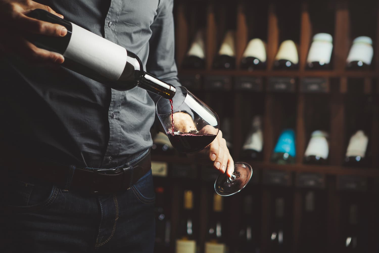 A person pours red wine from a bottle into a glass, with a wine rack in the background. A person pours red wine from a bottle into a glass, with a wine rack in the background.