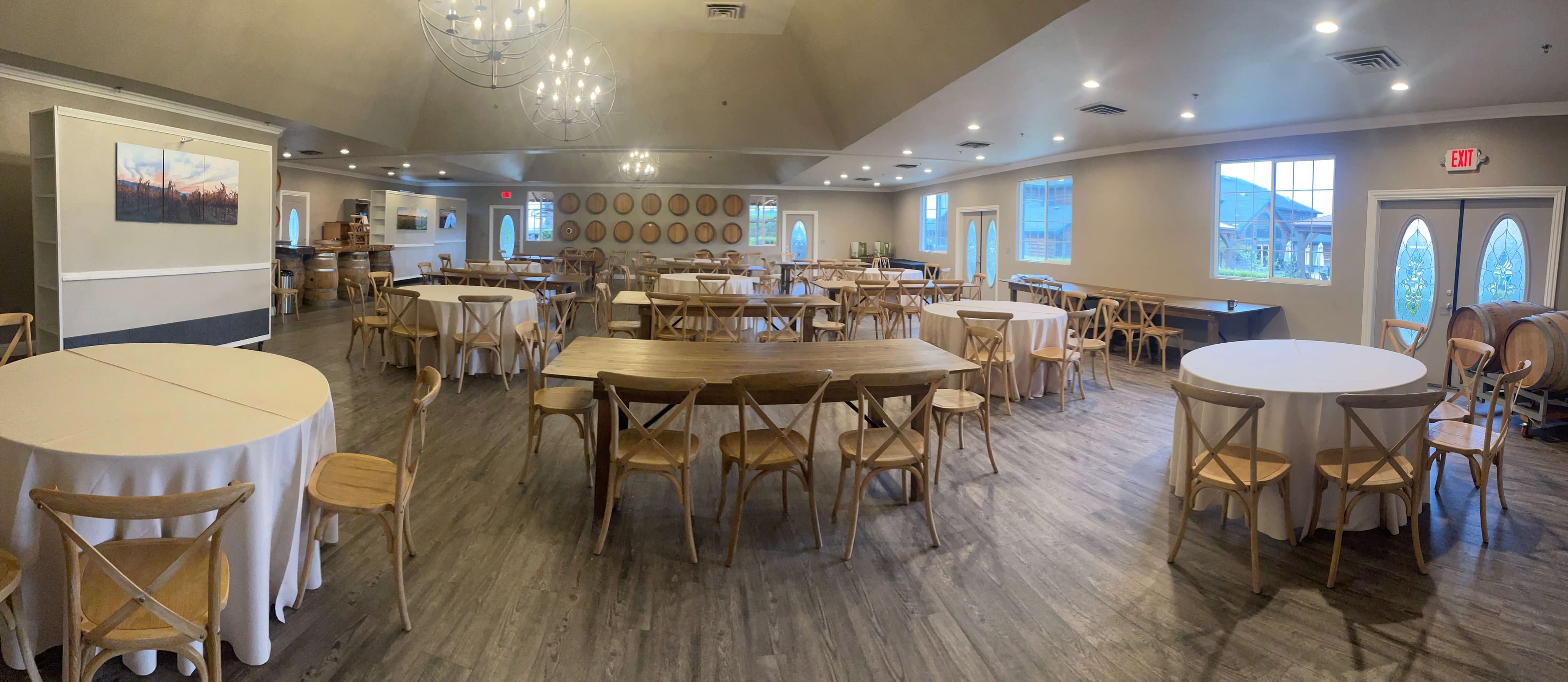 A spacious event room featuring round tables, wooden chairs, and large windows, with decorative wine barrels in the background.