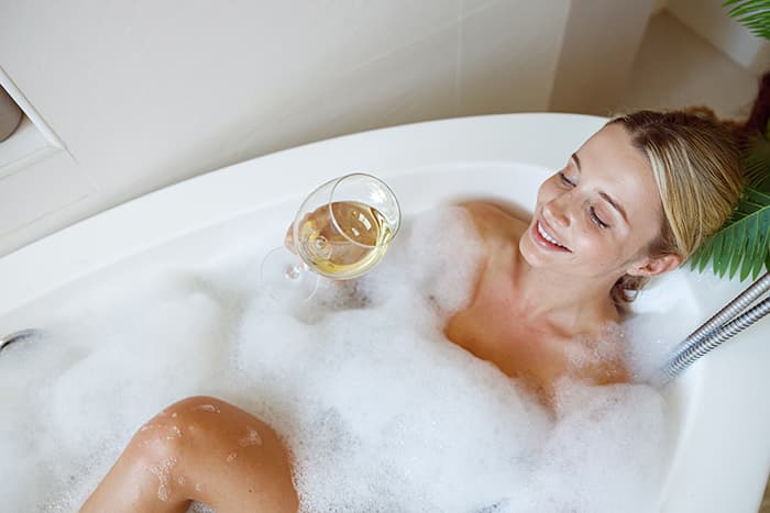 A woman relaxes in a bubble-filled bathtub while holding a glass of white wine.