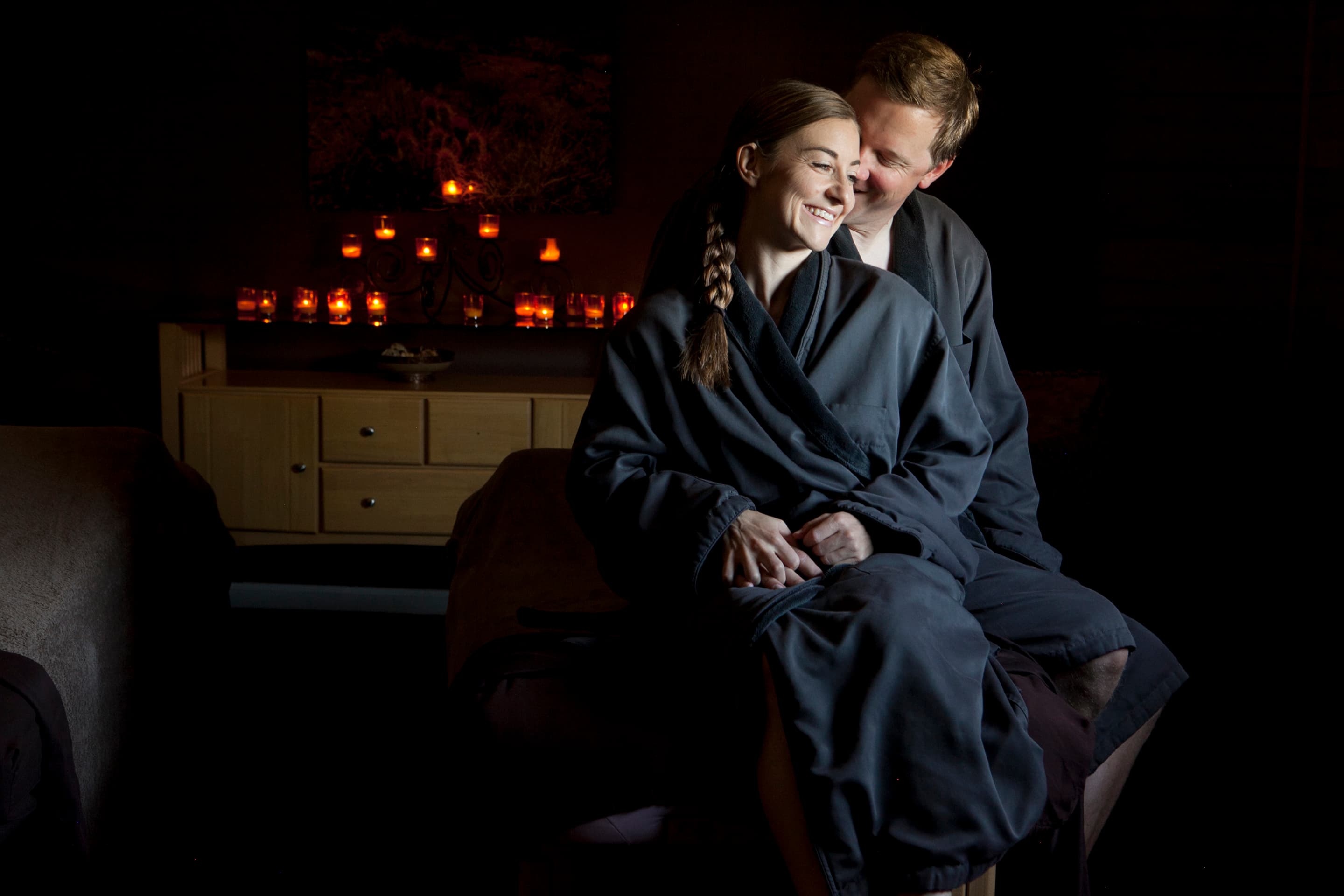 A couple in matching robes smiles at each other in a softly lit spa setting.