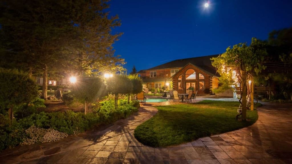 A beautifully landscaped backyard features a well-lit patio and a cozy log cabin under a full moon.