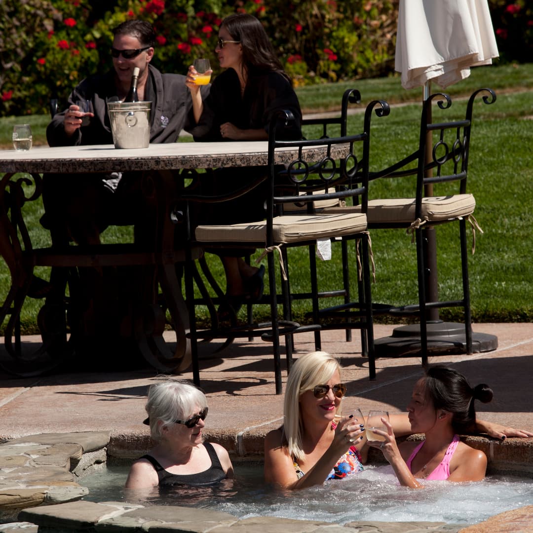 Two women enjoy drinks in a hot tub while two others relax at a nearby table.