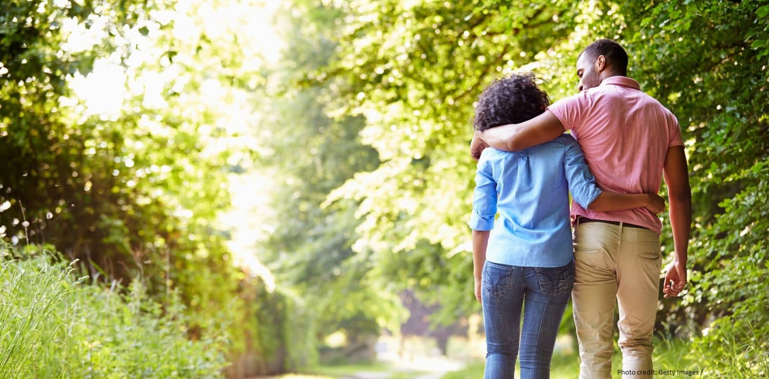 A couple strolls along a lush, green path, embracing each other.
