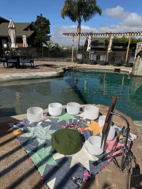 A peaceful outdoor setting featuring a quilt on the ground with crystal singing bowls and a meditative cushion beside a tranquil pool.