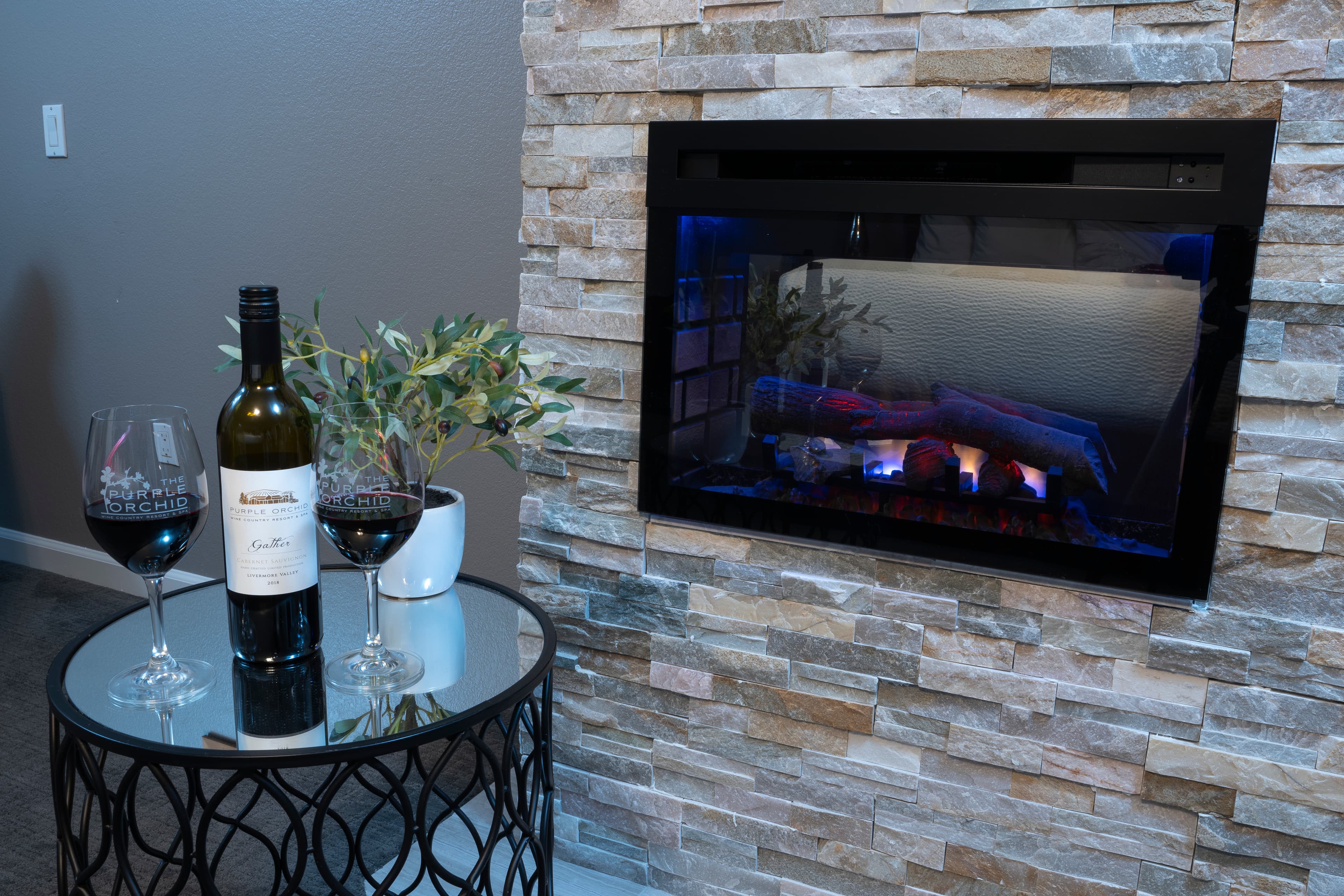 A tabletop with two wine glasses and a bottle sits in front of a modern electric fireplace.