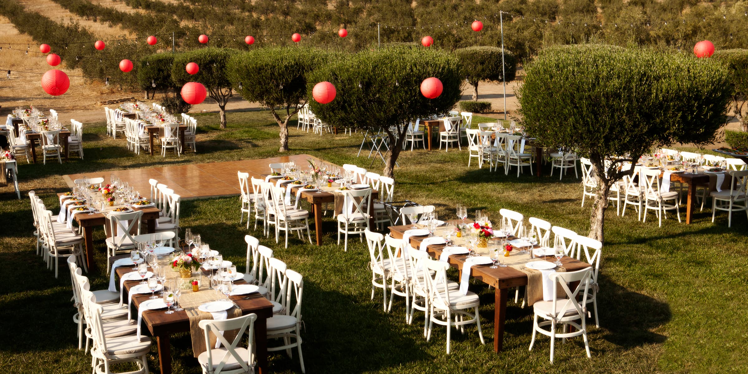 An outdoor dining setup with wooden tables and white chairs, decorated with red lanterns and surrounded by greenery.