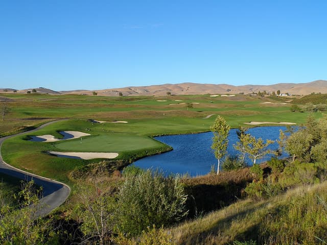 A panoramic view of a golf course with rolling hills, a lake, and clear blue skies.