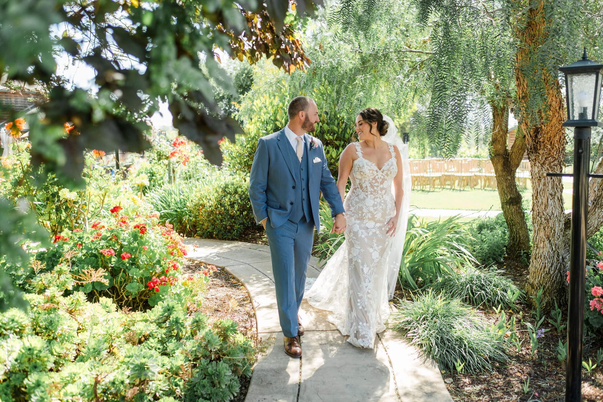 A bride and groom walk hand in hand along a flower-lined path in a sunny garden.