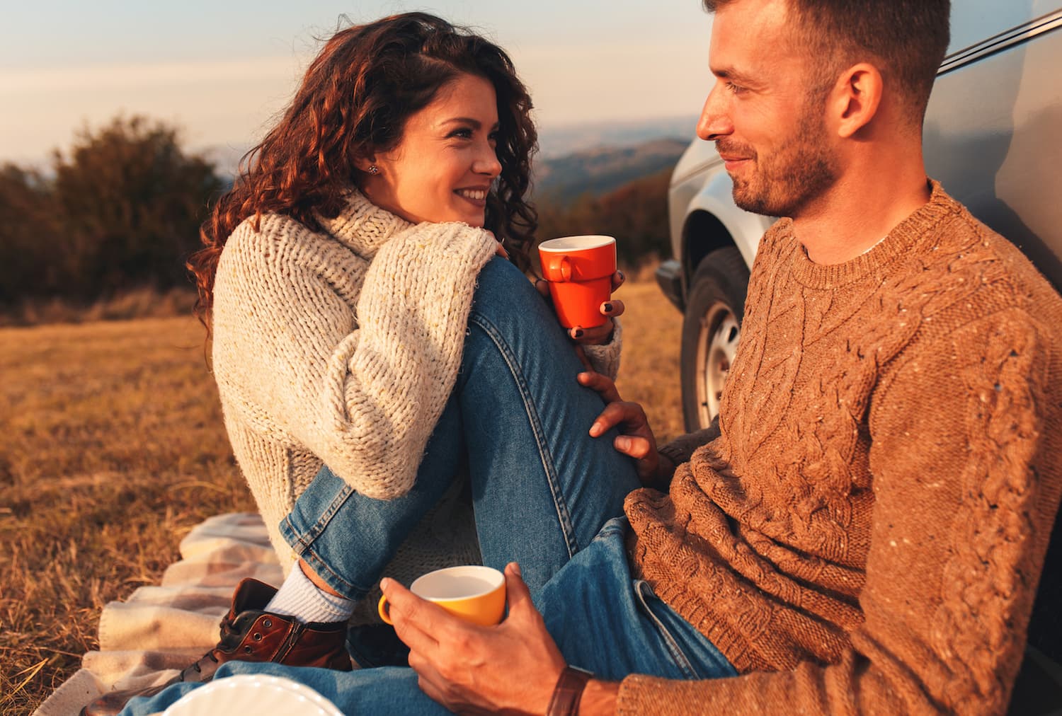 A smiling couple sits on a blanket outdoors, sharing mugs and enjoying each other's company. A smiling couple sits on a blanket outdoors, sharing mugs and enjoying each other's company.