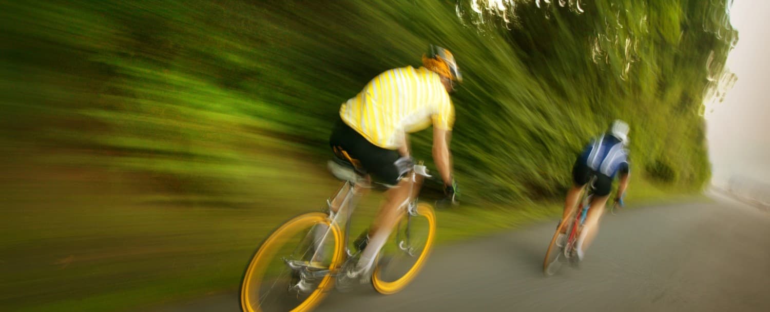 Two cyclists ride rapidly along a tree-lined path. Two cyclists ride rapidly along a tree-lined path.