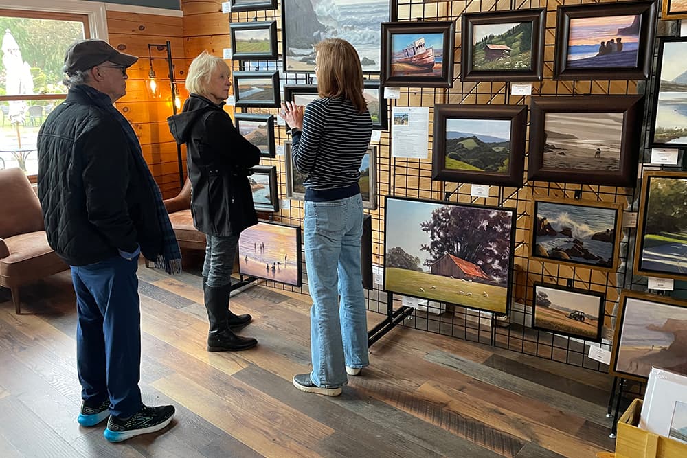 Three people discuss artwork displayed on a wall in an art gallery.