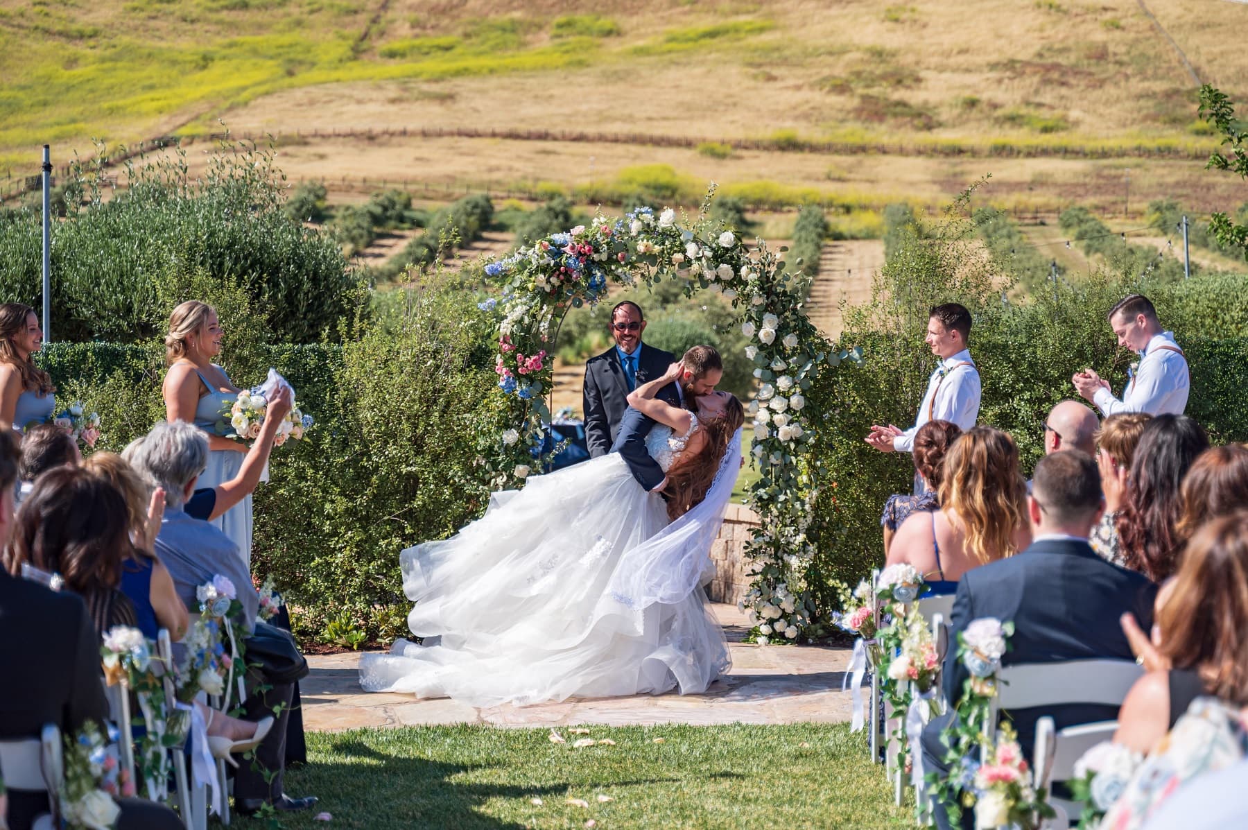 A couple shares a romantic embrace during their wedding ceremony, surrounded by guests and a floral arch.