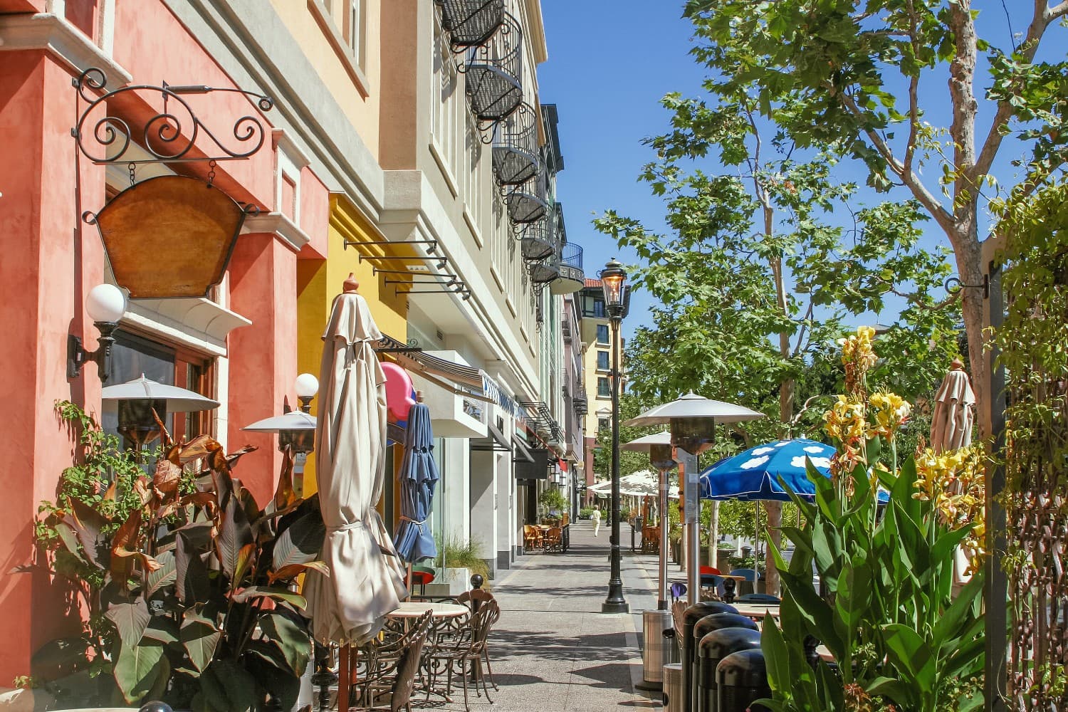 A lively street lined with cafes, umbrellas, and greenery under a clear blue sky.