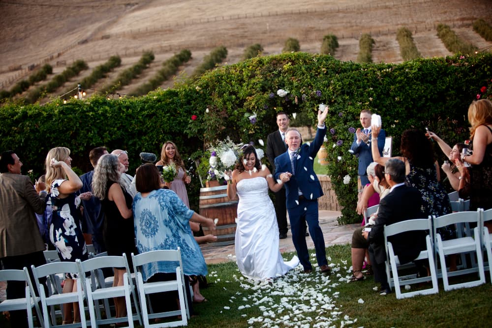 Bride and groom in vineyard with guests and flower petals flowing in air