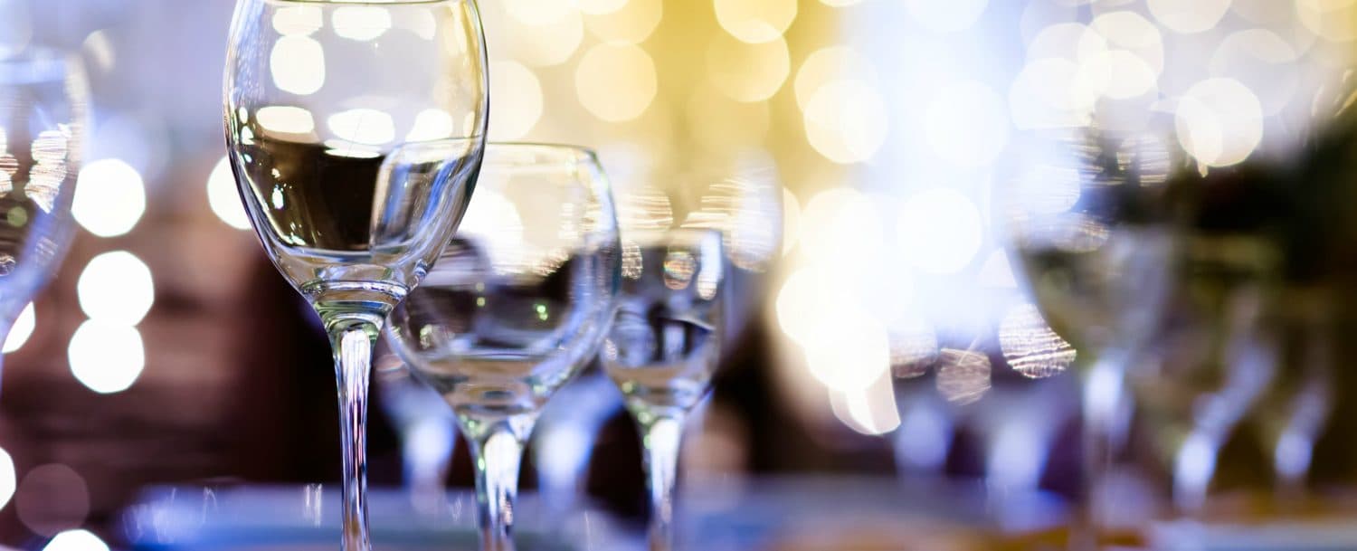 A row of empty wine glasses glistens in front of a softly lit, blurred background. A row of empty wine glasses glistens in front of a softly lit, blurred background.