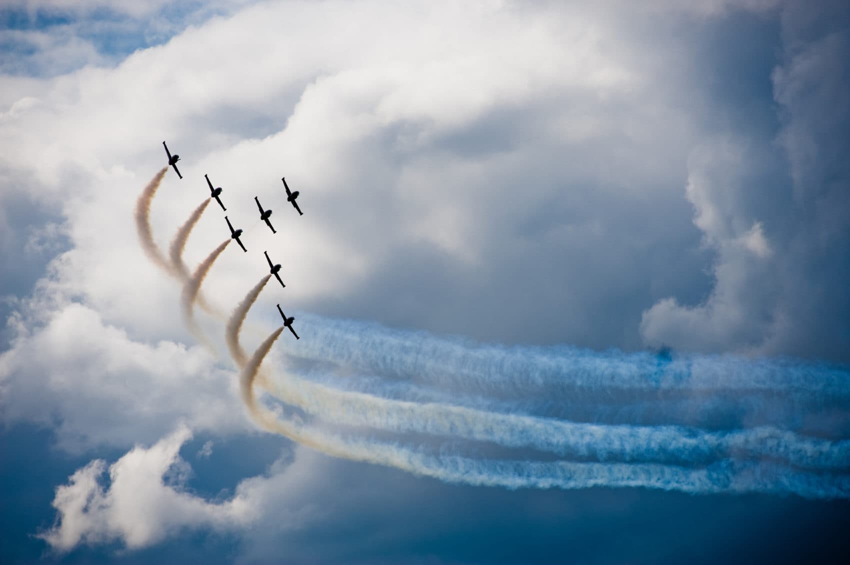 A formation of six planes performs aerial maneuvers, leaving colorful smoke trails against a cloudy sky. A formation of six planes performs aerial maneuvers, leaving colorful smoke trails against a cloudy sky.