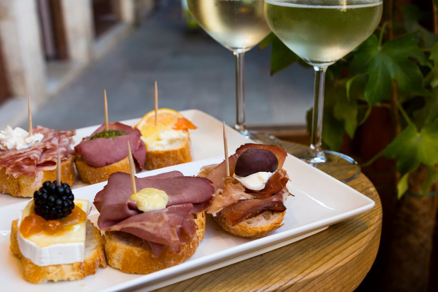 A platter of assorted bruschetta topped with meats and cheeses, accompanied by two glasses of white wine. A platter of assorted bruschetta topped with meats and cheeses, accompanied by two glasses of white wine.