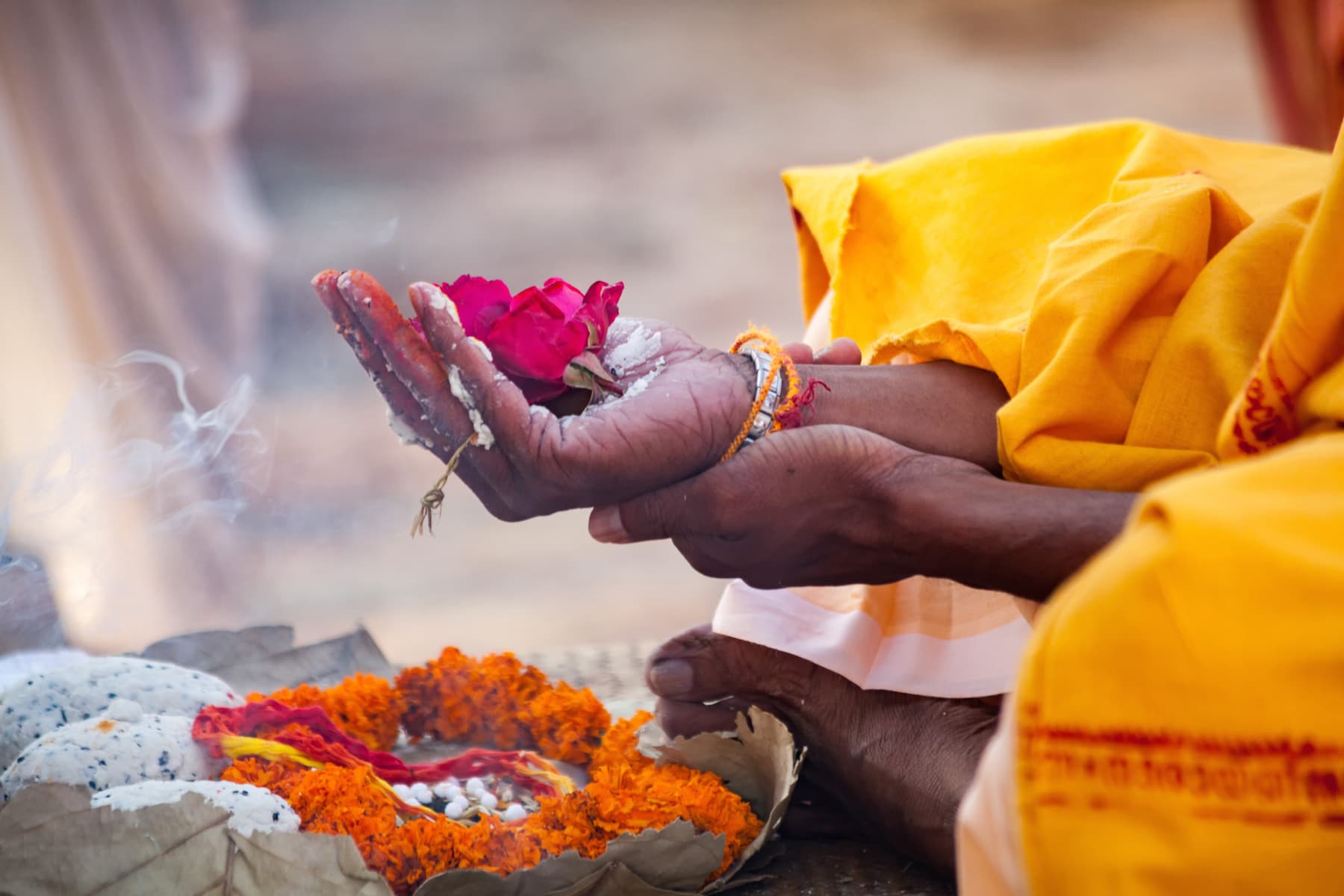A close-up of hands holding a rose and surrounded by colorful flower offerings and smoke.