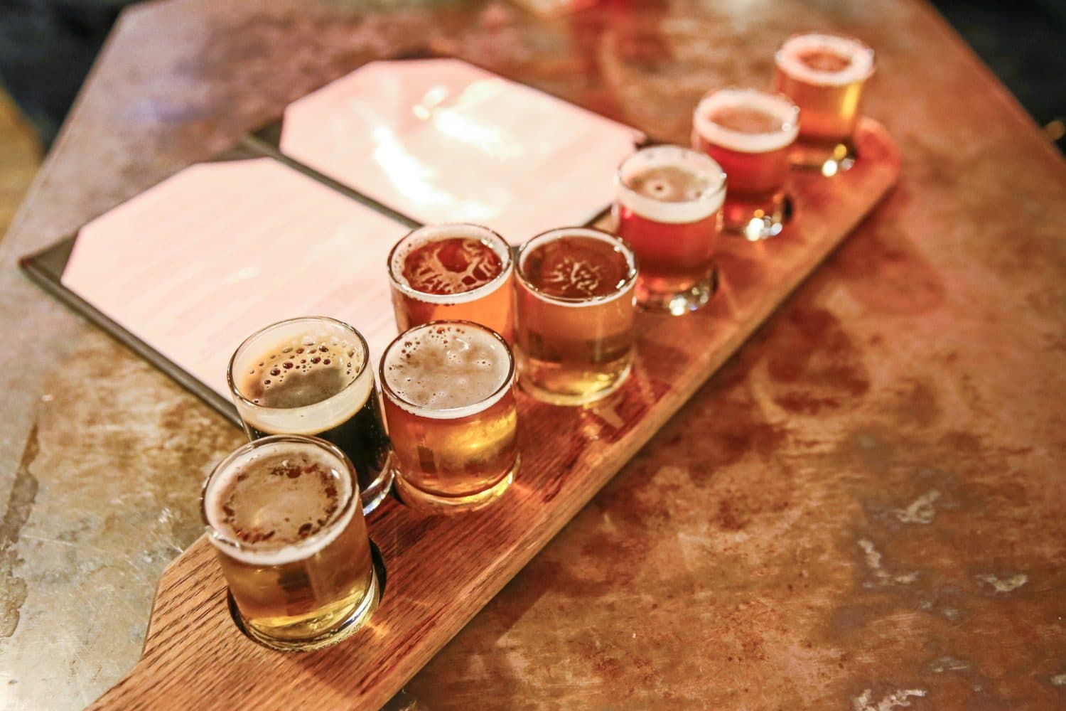 A wooden flight tray holds eight small glasses of various beers next to a menu.