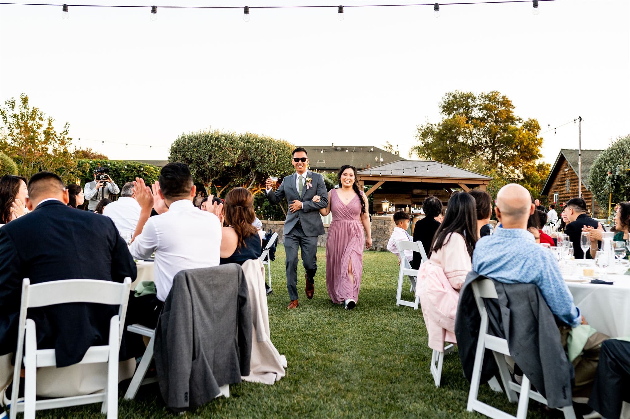 A man and woman joyfully enter a garden venue, greeted by applauding guests seated at tables.