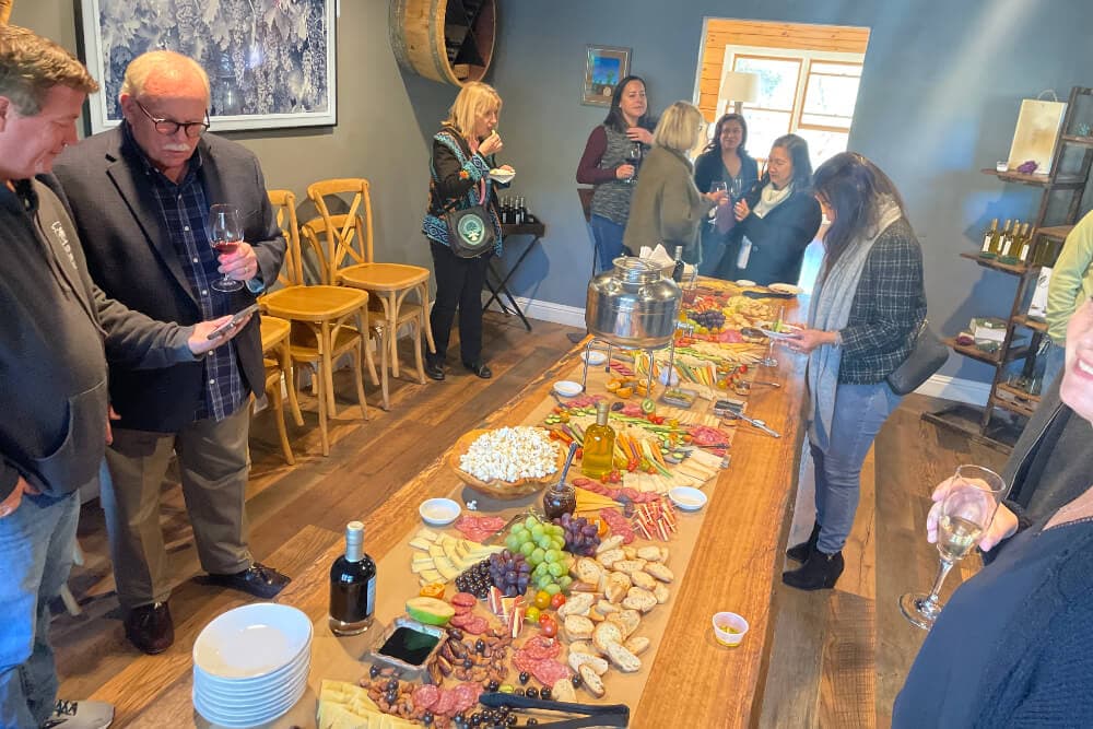 A group of people mingles around a large charcuterie spread in a cozy setting.